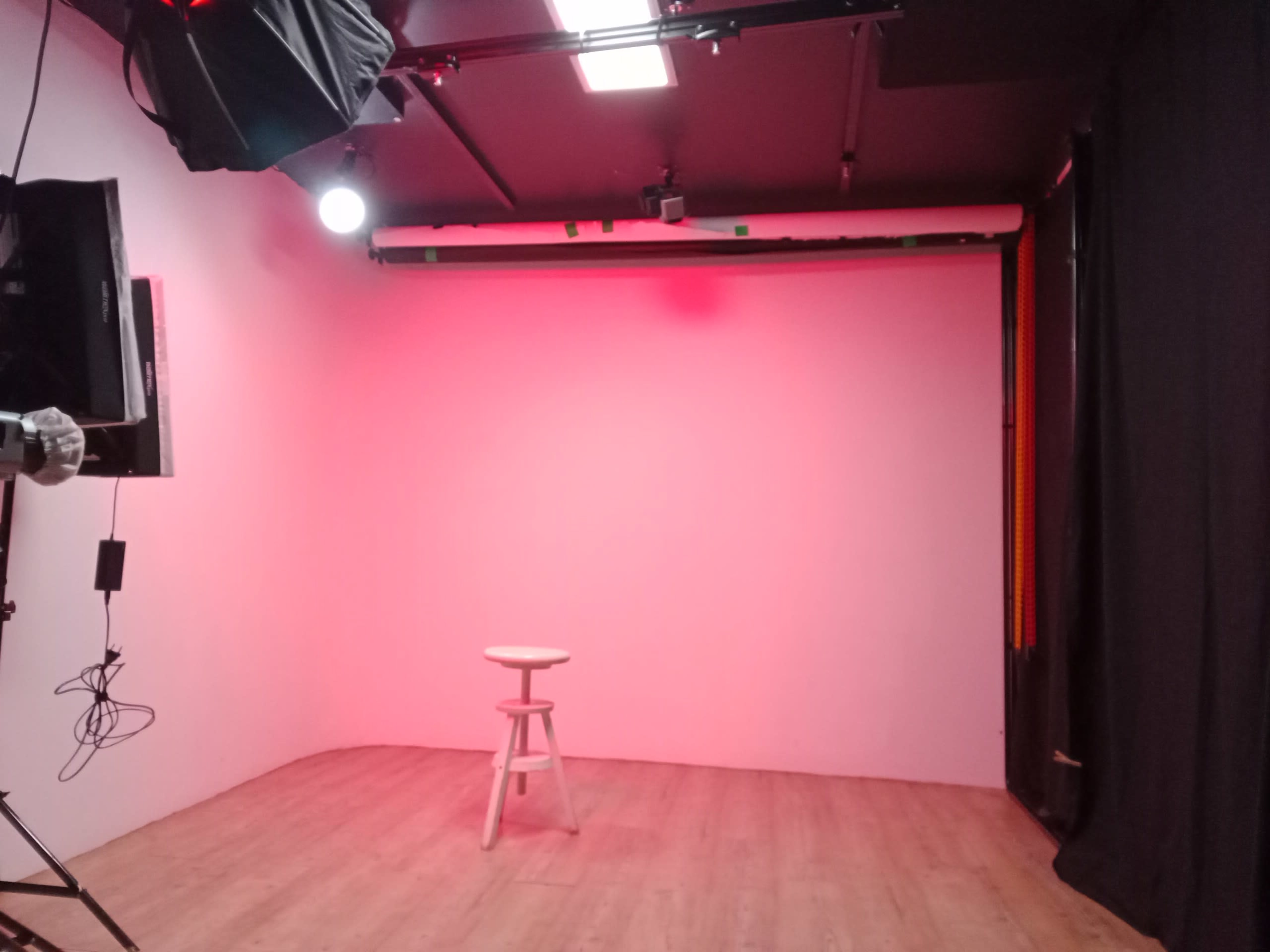 The image shows an empty photo studio with a wooden stool positioned in front of a pink backdrop, illuminated by studio lights.