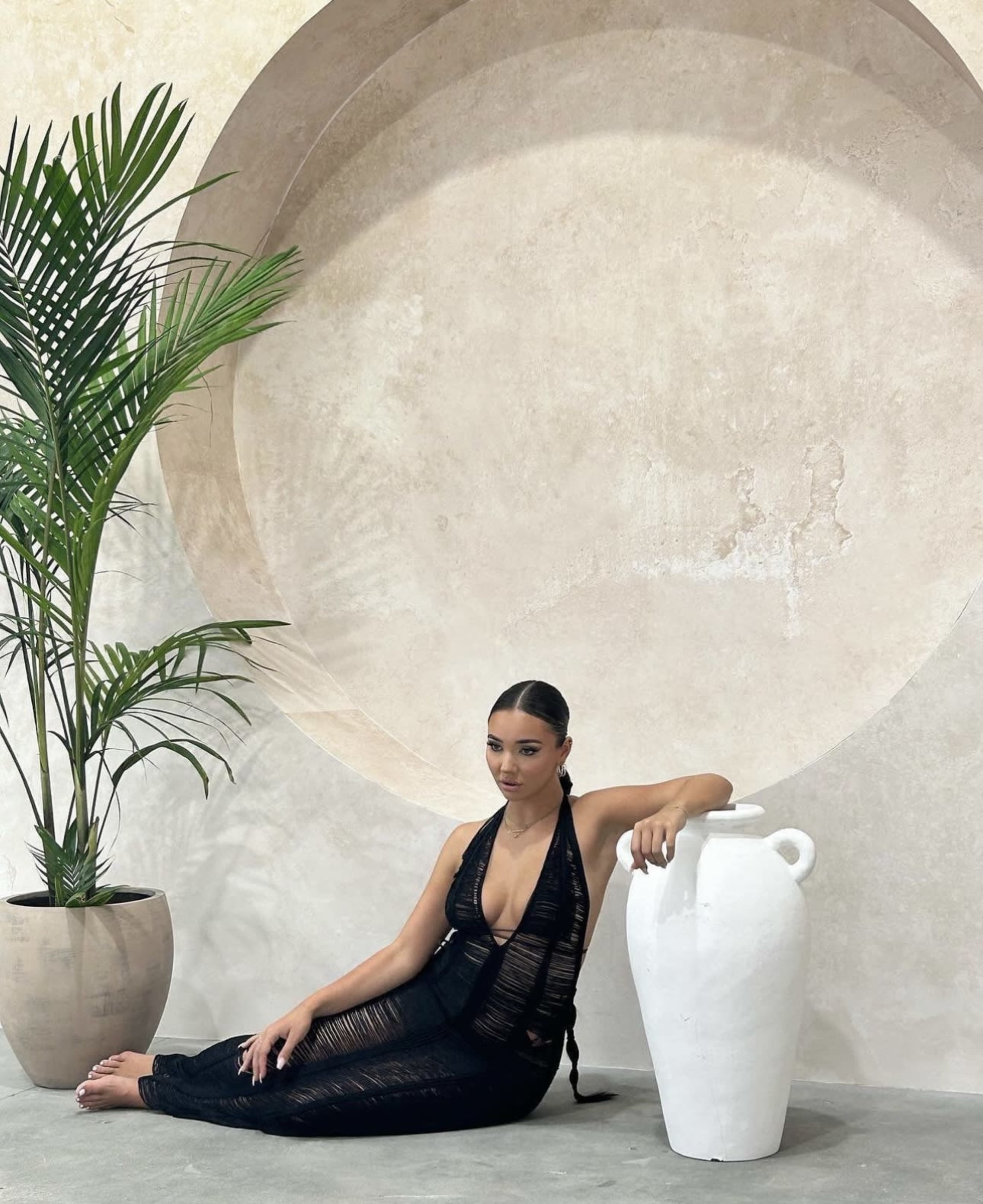 A woman sits beside a large white vase in front of a circular wall, with a potted palm plant nearby.