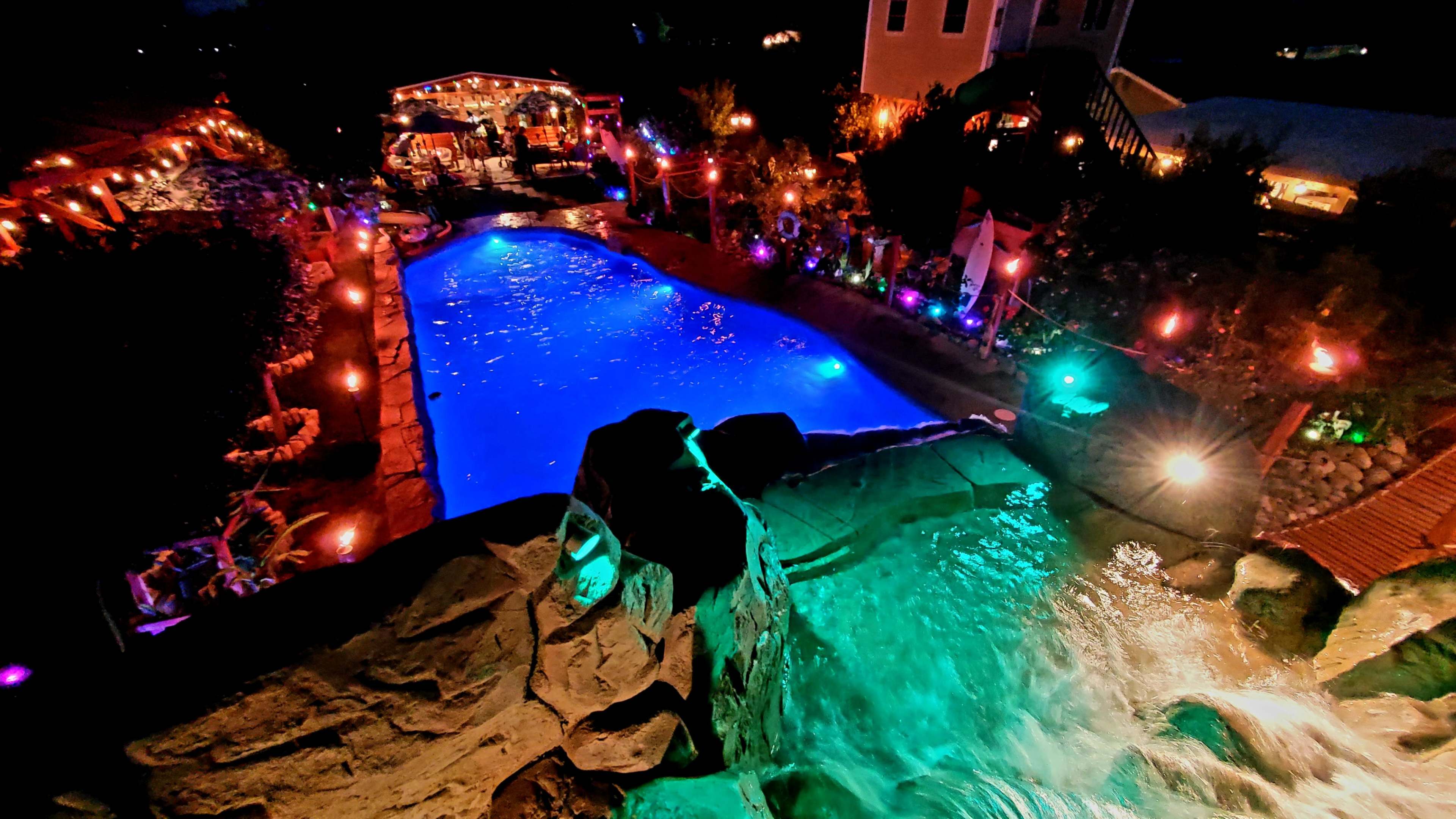 The image shows a lit outdoor pool area with colorful lights, a waterfall feature, and a gathering space in the background.