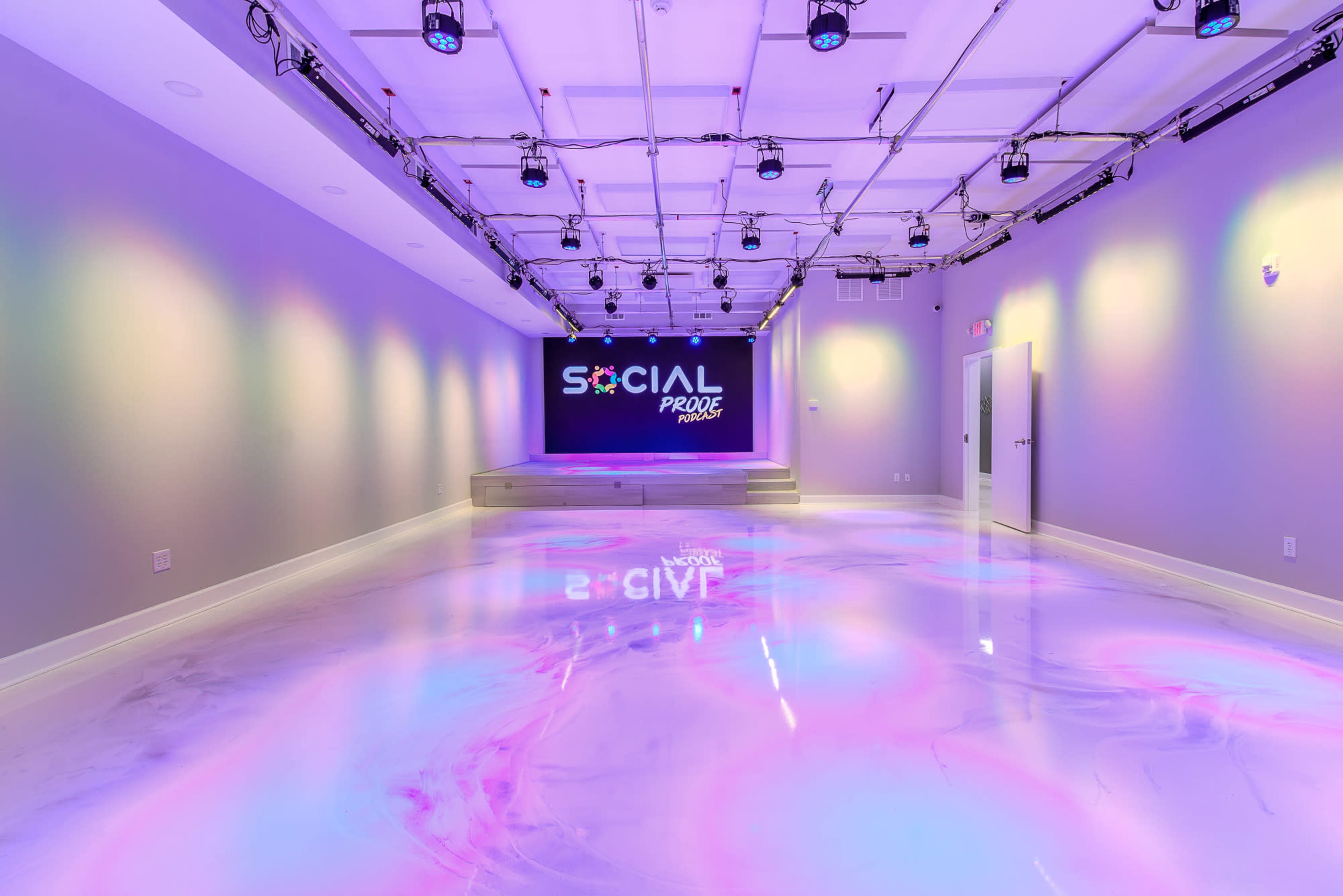 The image shows a modern event space with purple and pastel lighting, featuring a stage area and a glossy floor that reflects the vibrant colors.