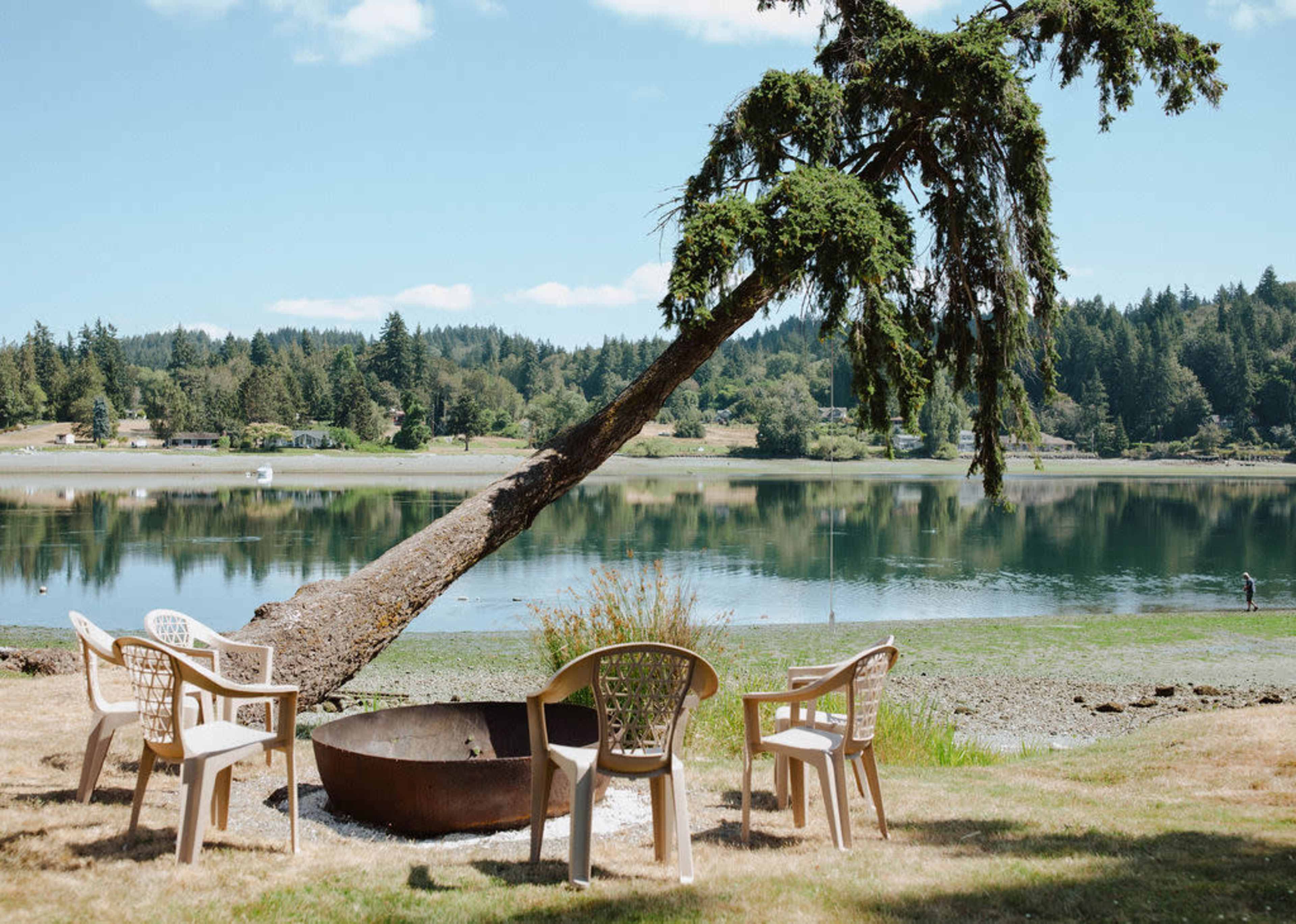 A fire pit surrounded by four plastic chairs is positioned by a calm lake with trees in the background.
