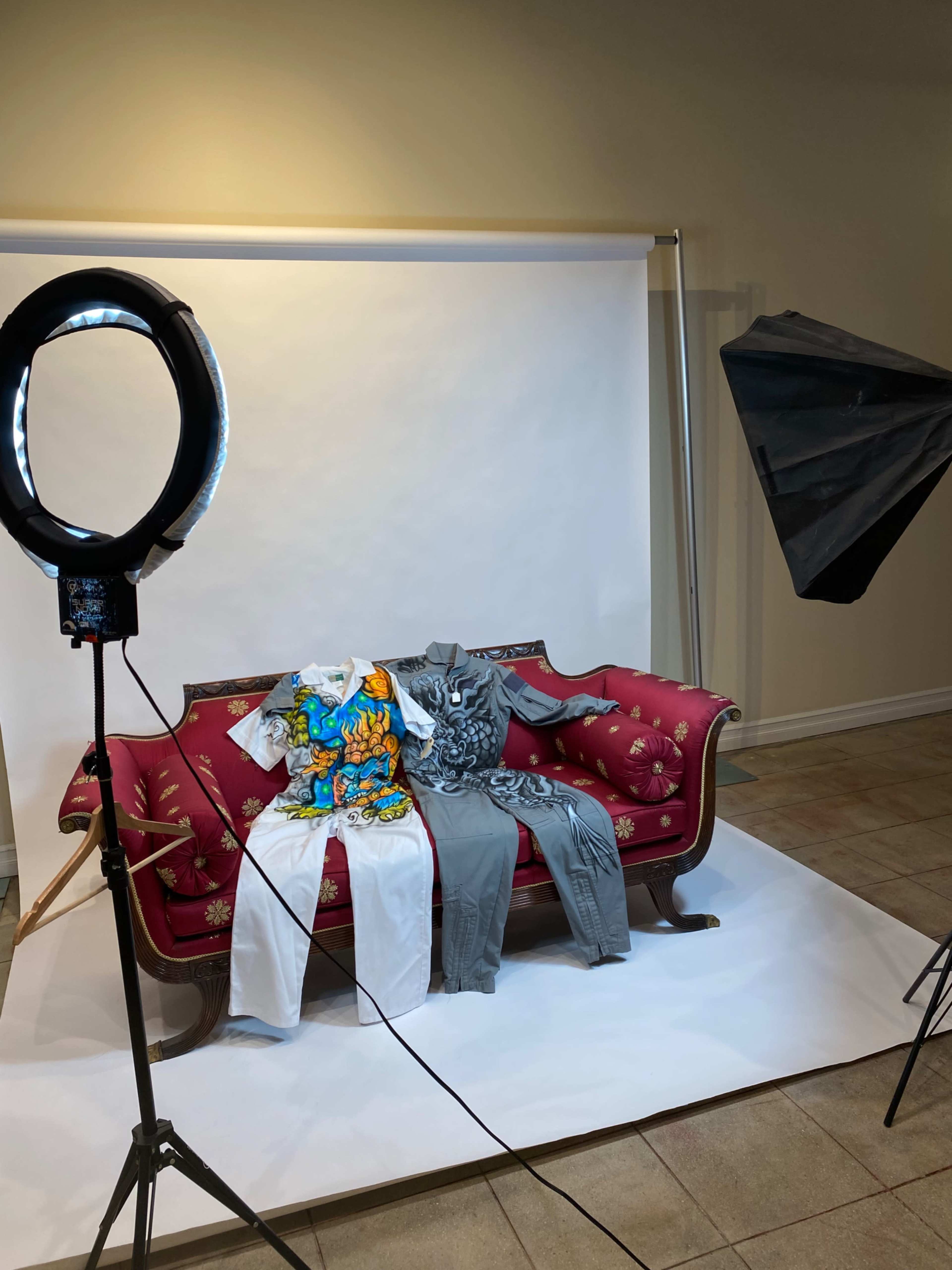A red sofa is positioned against a white backdrop with three outfits displayed on it, accompanied by studio lighting equipment on either side.