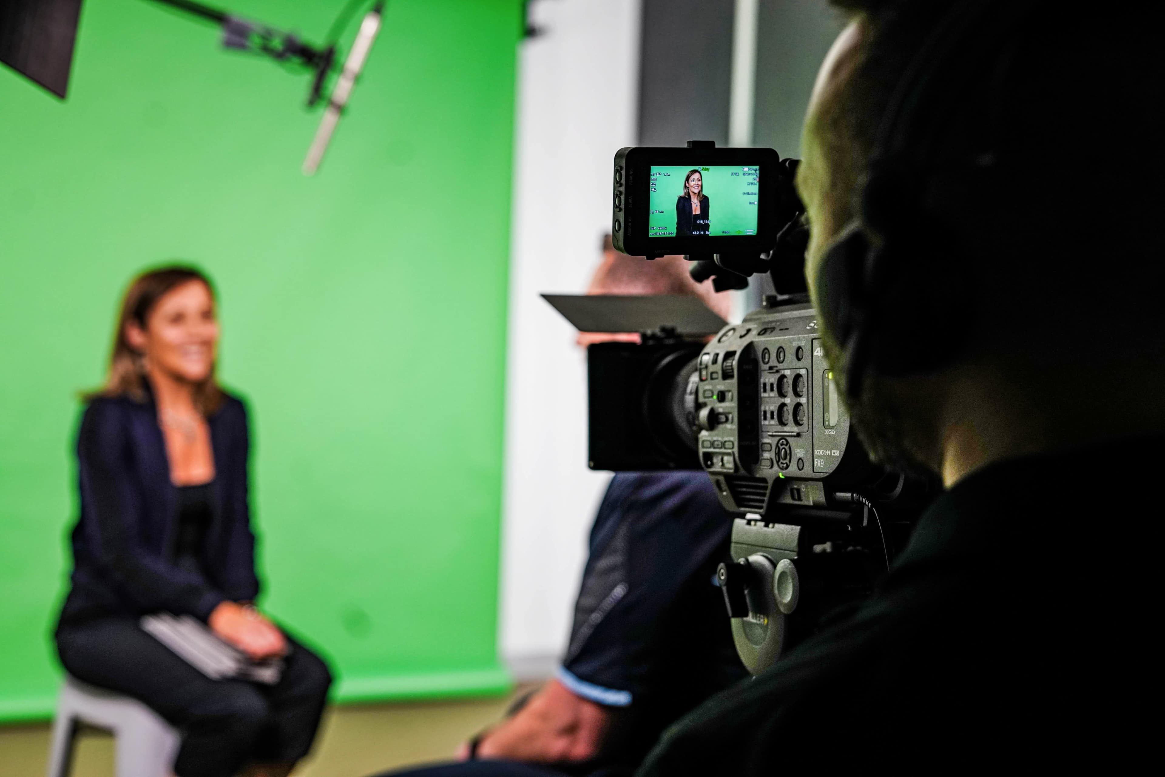 A woman is being filmed in front of a green screen while a camera operator monitors the shot.
