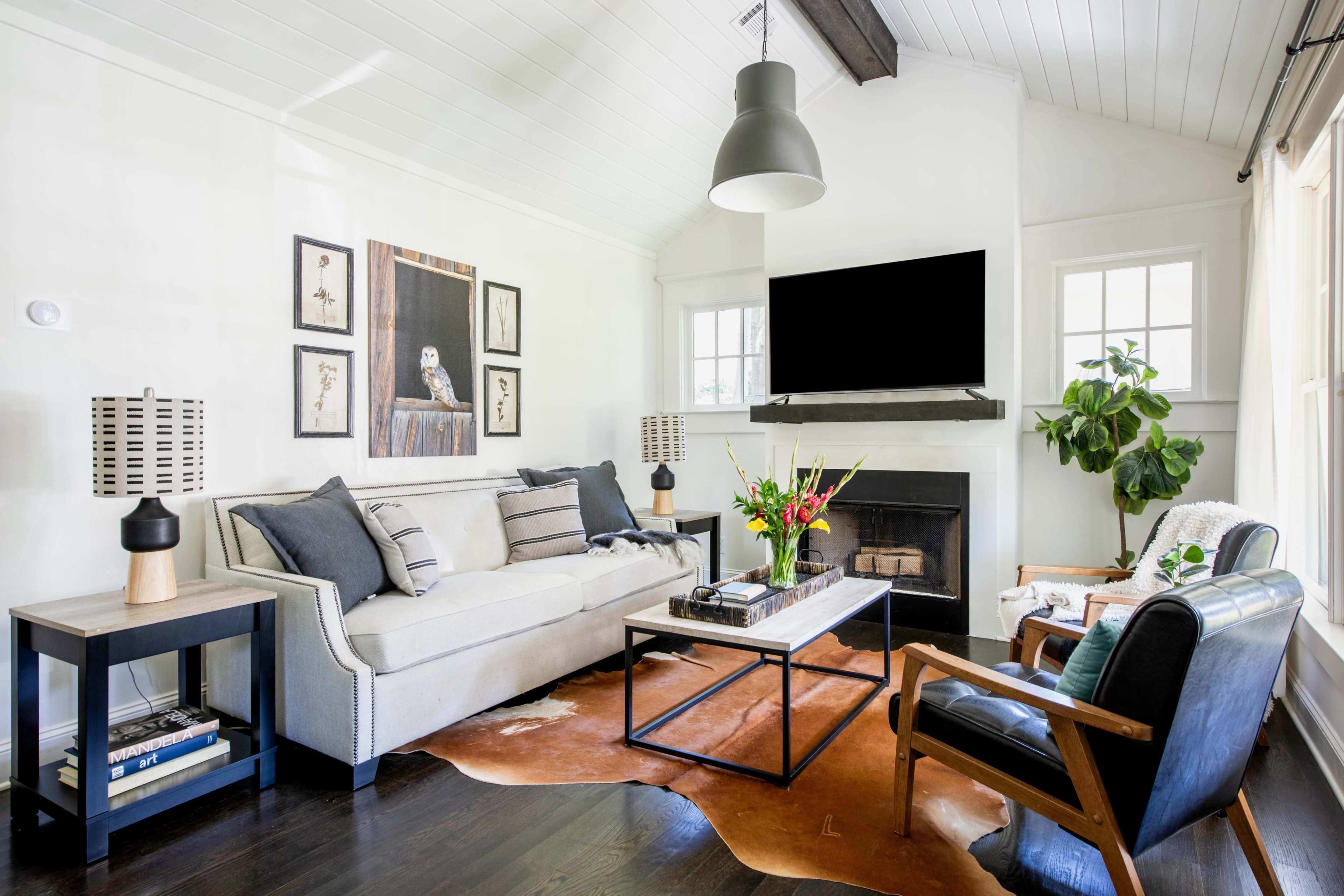 A modern living room featuring a beige sofa, a black coffee table, and a large television mounted above a fireplace, with accent chairs and decorative plants.