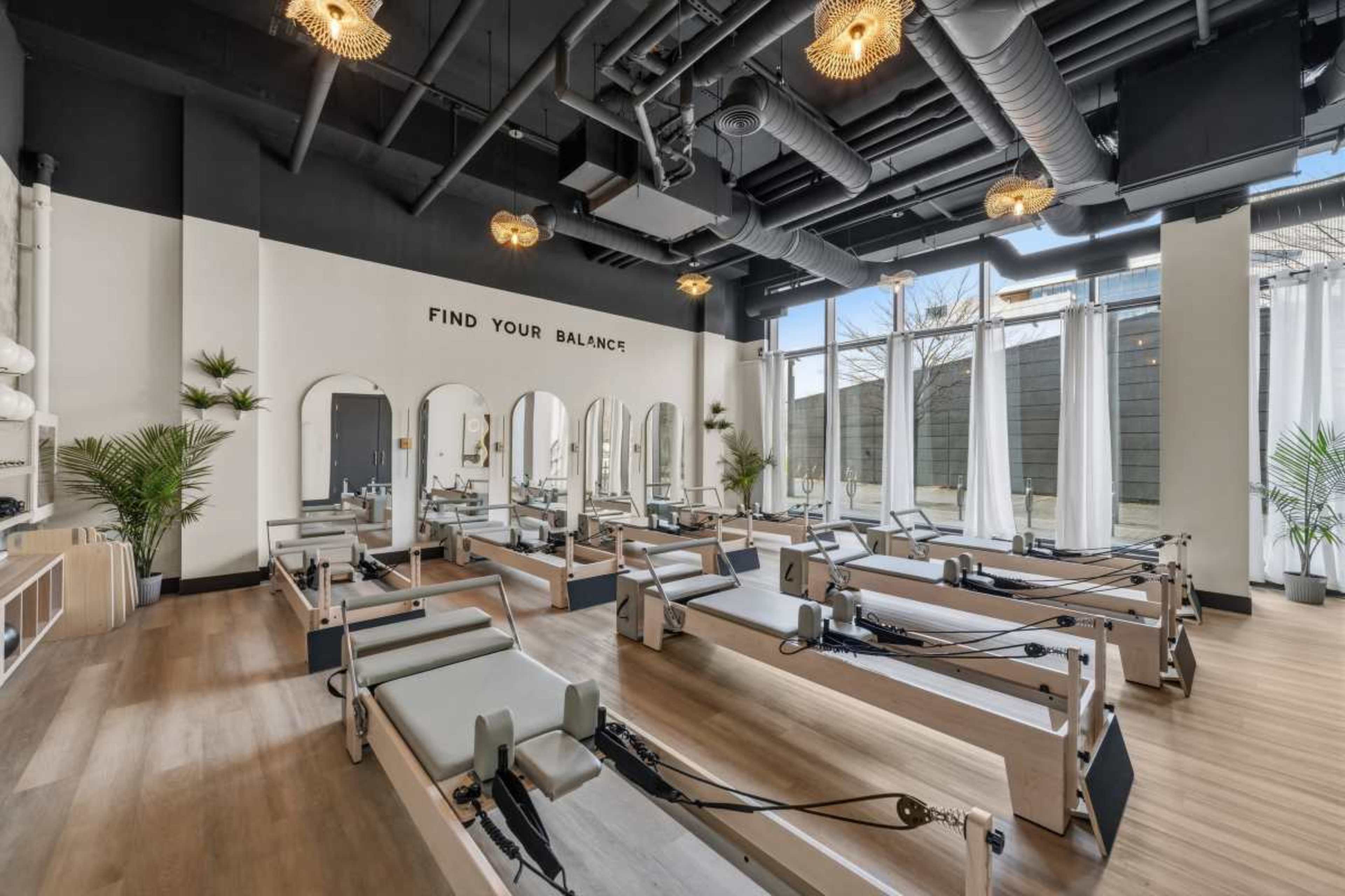 The image shows a modern pilates studio with rows of reformer machines, large windows letting in natural light, and minimalistic decor.