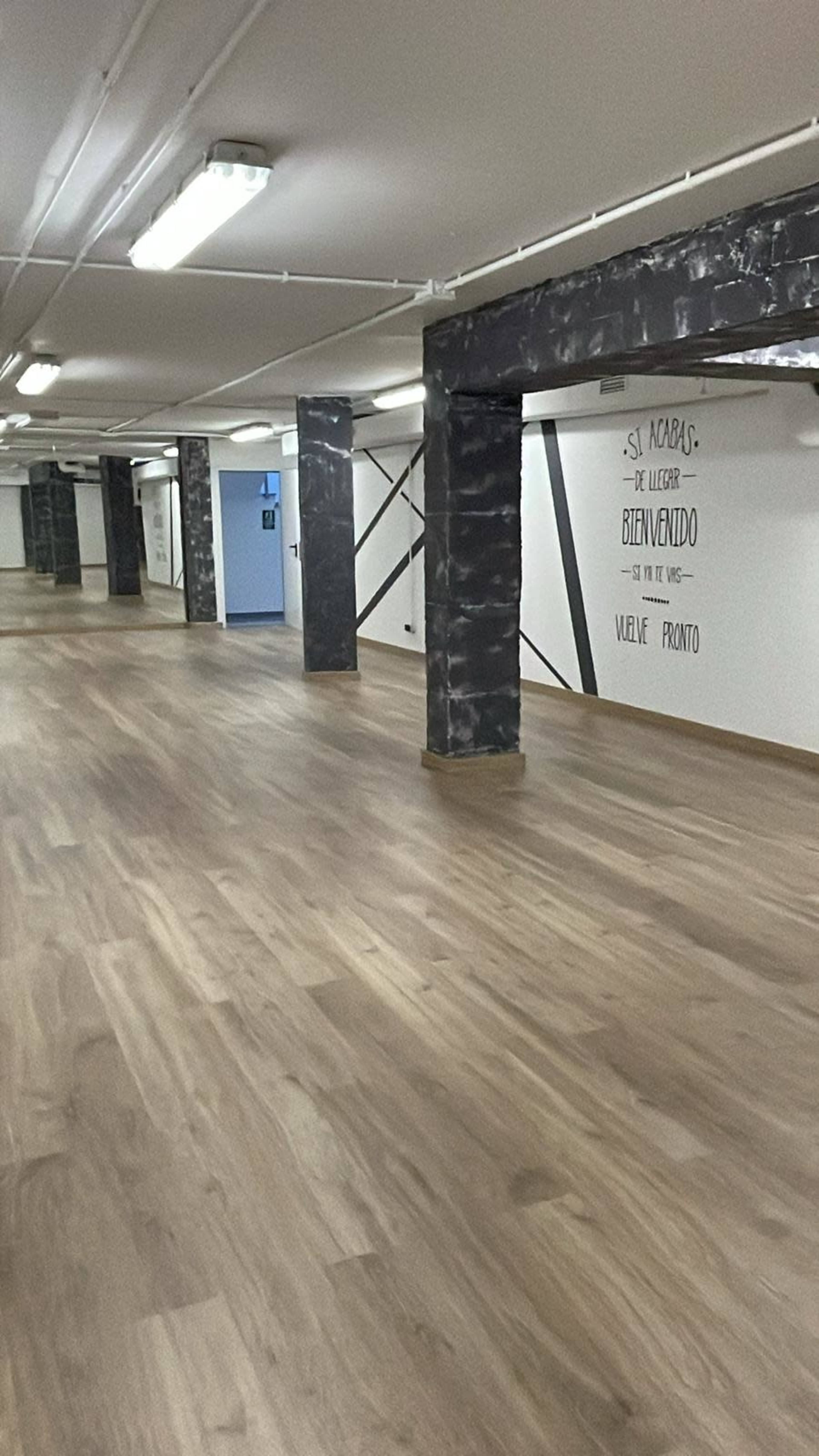 A spacious interior room with wooden flooring and concrete pillars, featuring a wall with text in Spanish.