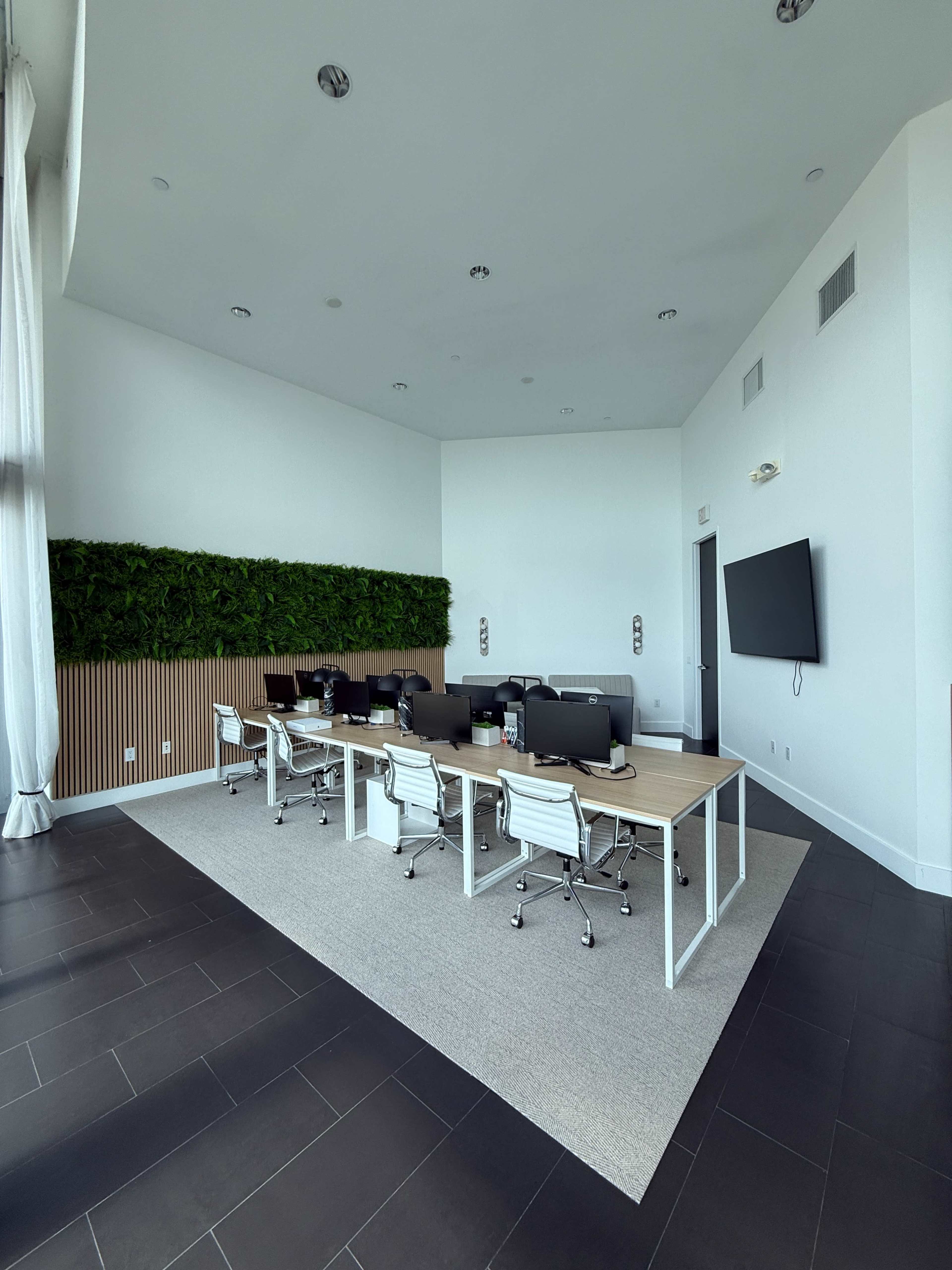 The image shows a modern office space featuring a row of desks with computer monitors, white ergonomic chairs, and a vertical green wall.