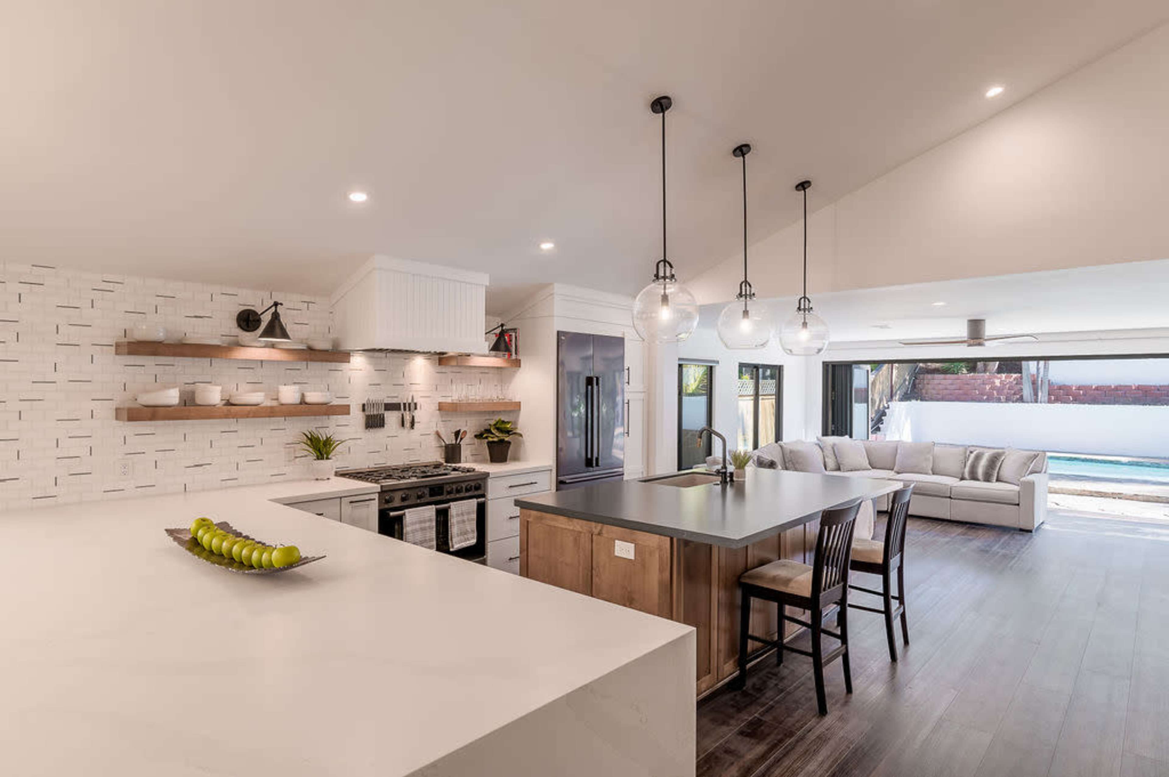 A modern kitchen features a large island, stainless steel appliances, and an open layout that connects to a living area with a sofa and a view of a pool.