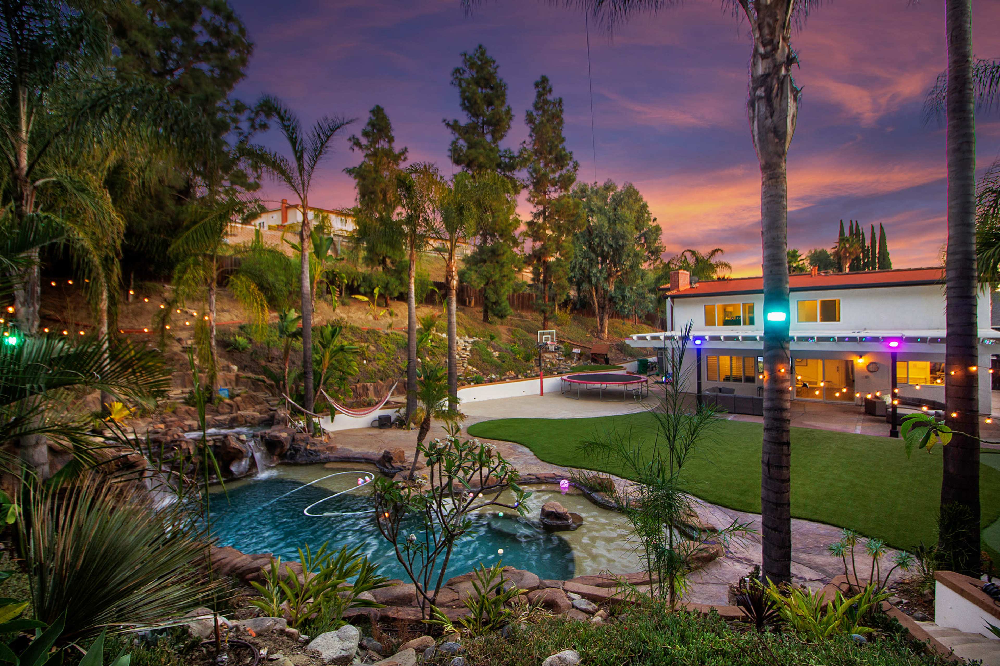 A backyard landscape featuring a swimming pool, landscaped greenery, and a house with outdoor lighting against a sunset sky.