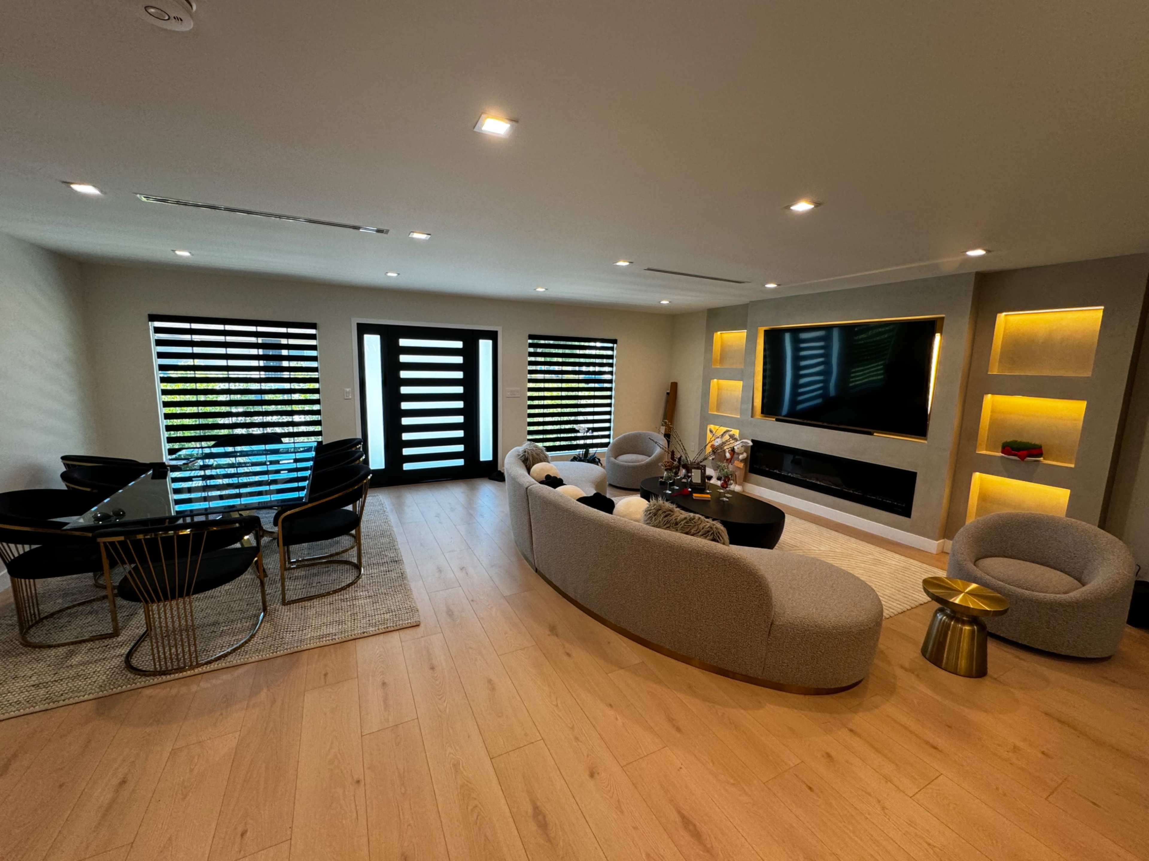 A modern living room with a curved sofa, accent chairs, a black coffee table, and a dining area featuring a glass table and black chairs, illuminated by built-in lighting.