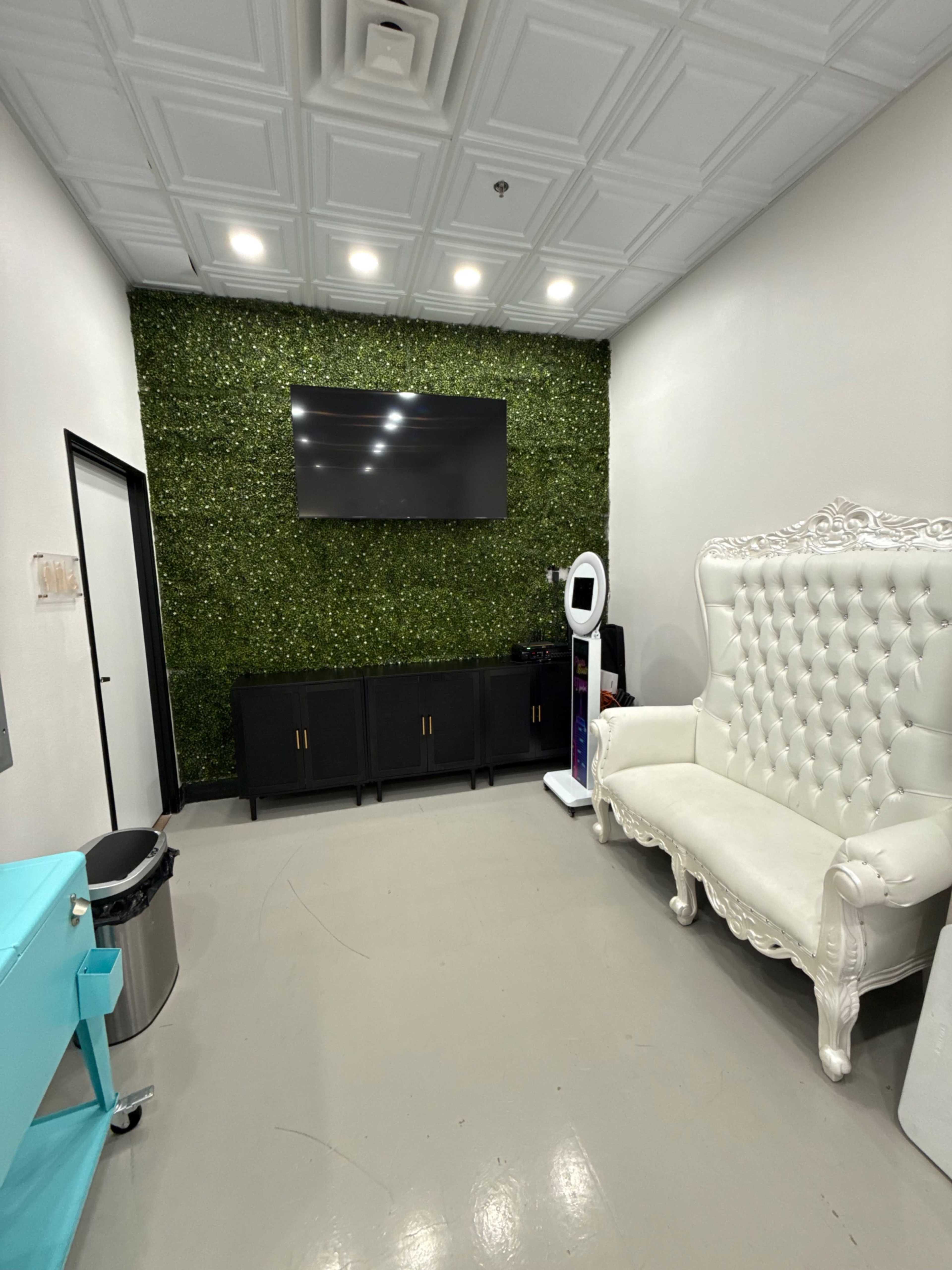 The image shows a small room with a green wall covered in artificial foliage, a large white chair, a black cabinet, a television mounted on the wall, and a kiosk in the corner.
