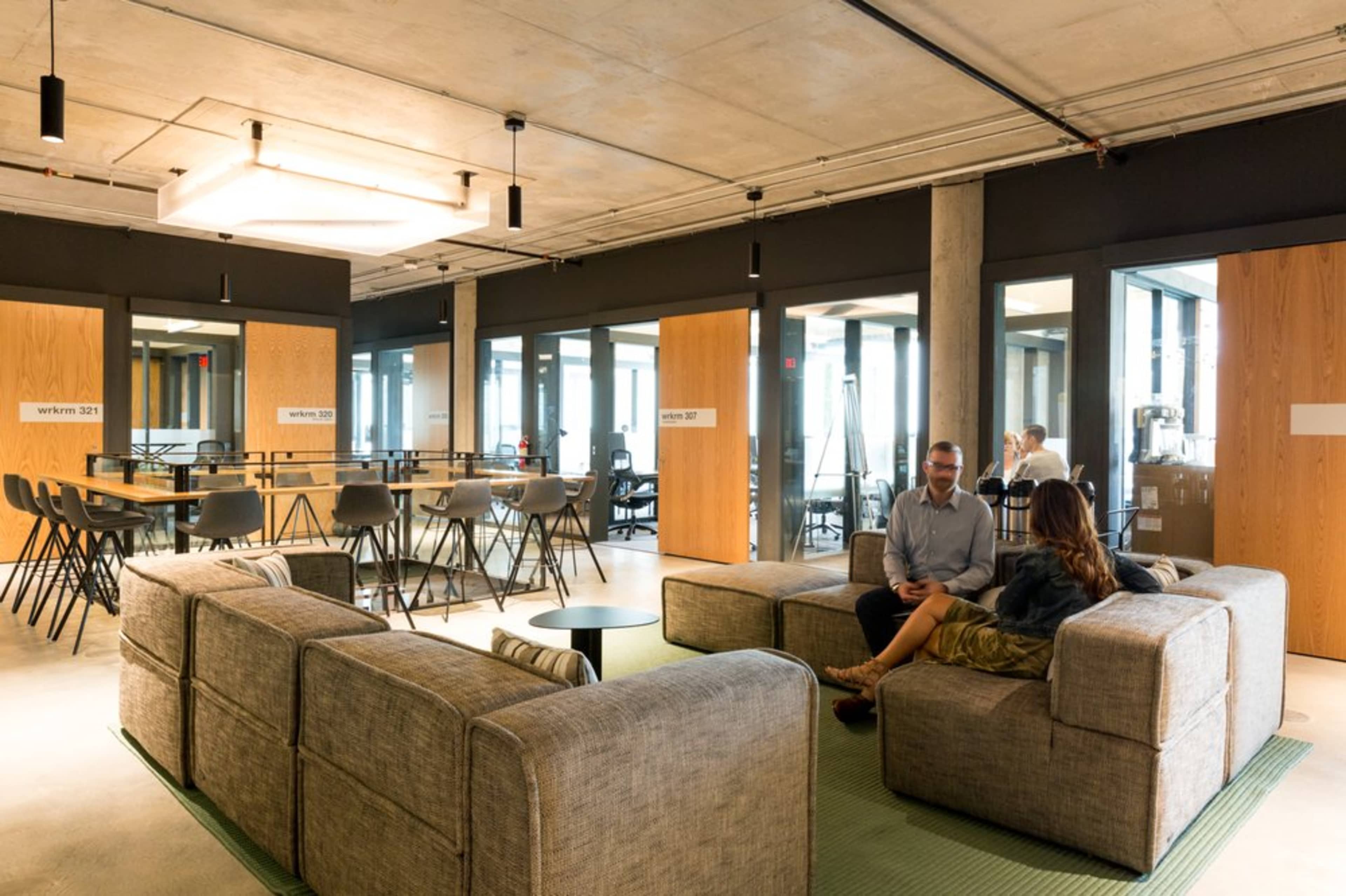 A modern office space with a seating area composed of gray couches and a coffee table, adjacent to glass-walled meeting rooms.