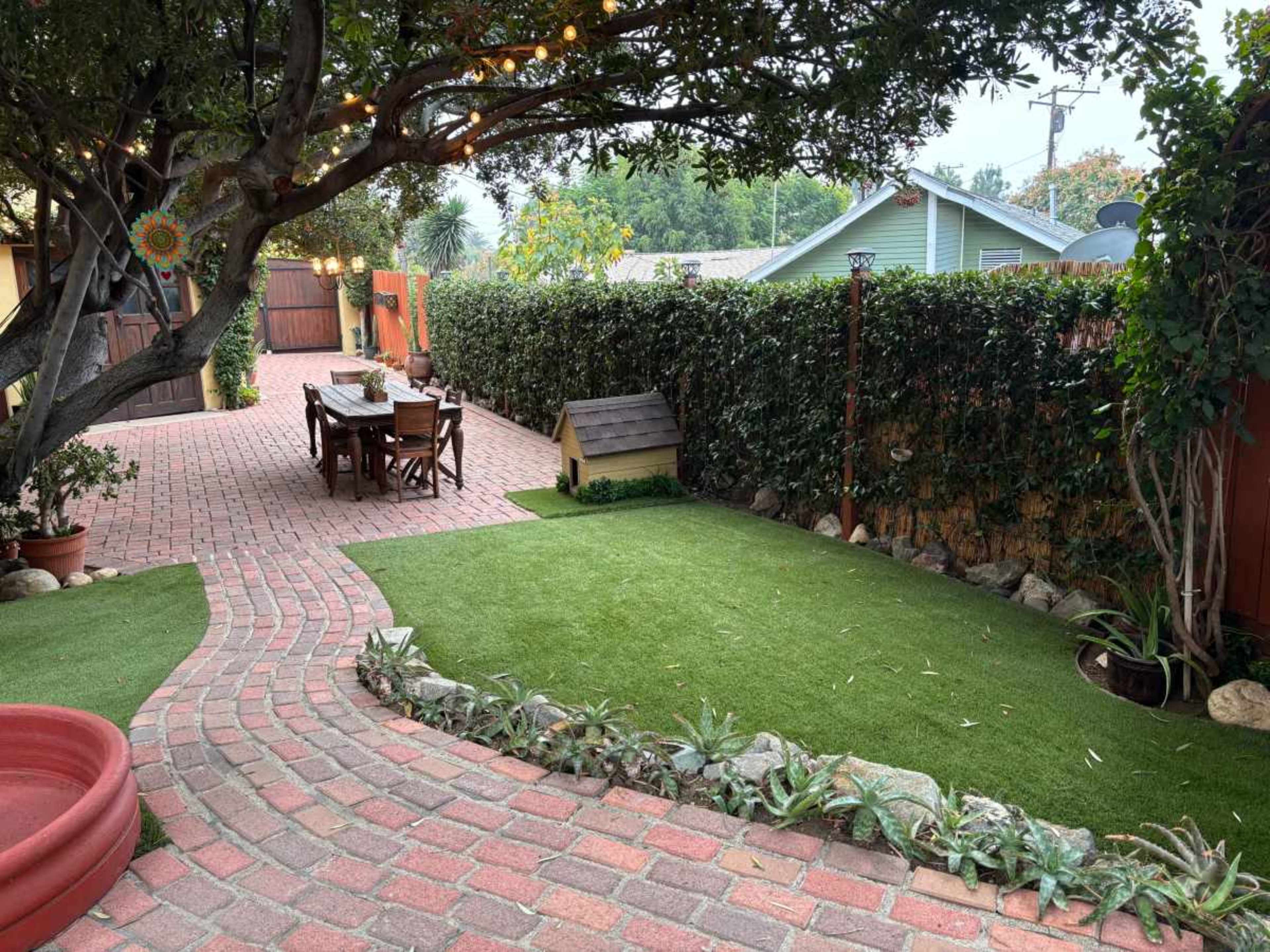 The image shows a brick patio with a wooden dining table and chairs, surrounded by greenery and a small doghouse.
