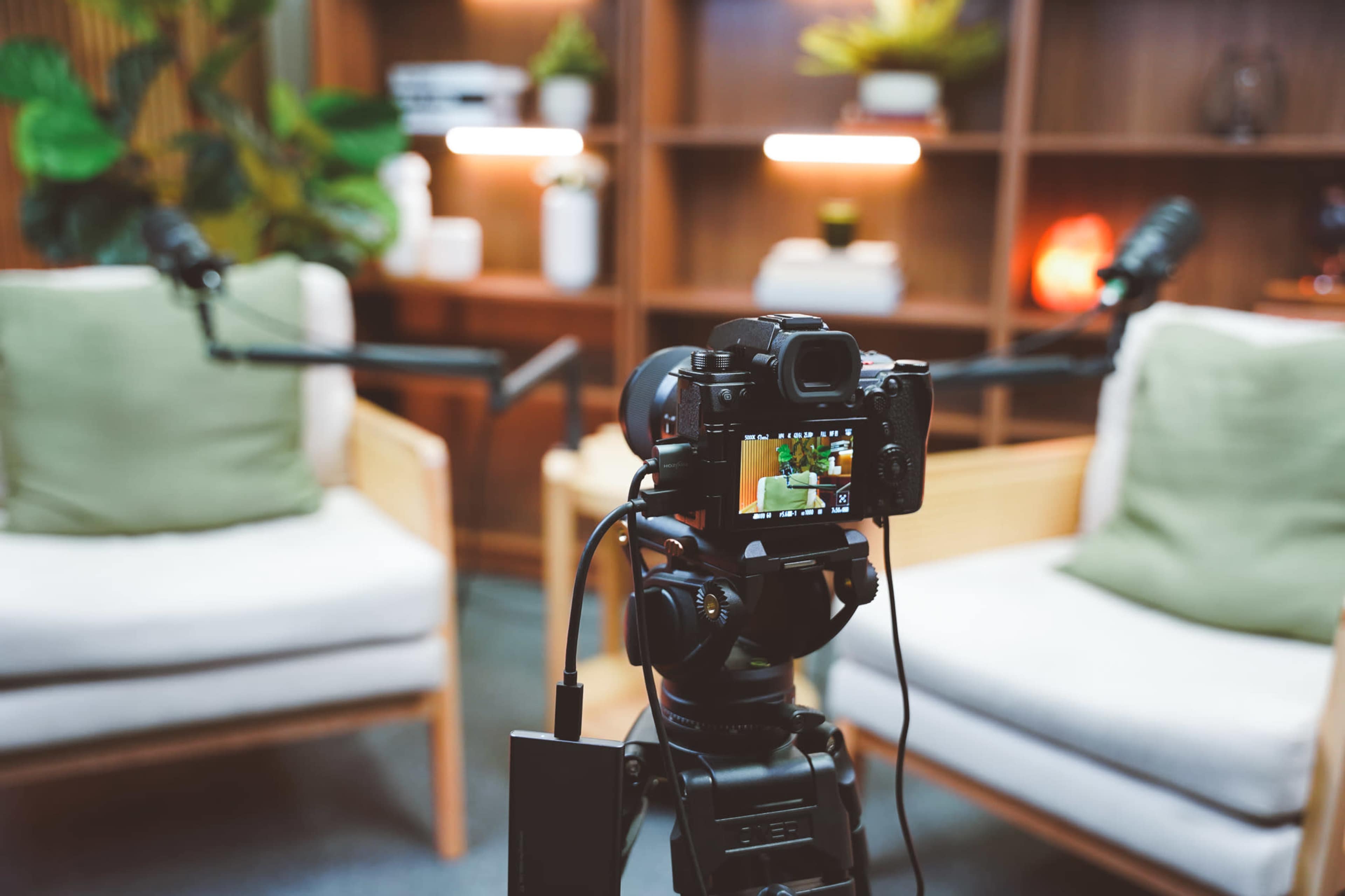 A camera is positioned in front of two upholstered chairs with microphones attached, set in a well-lit room with plants and shelves in the background.