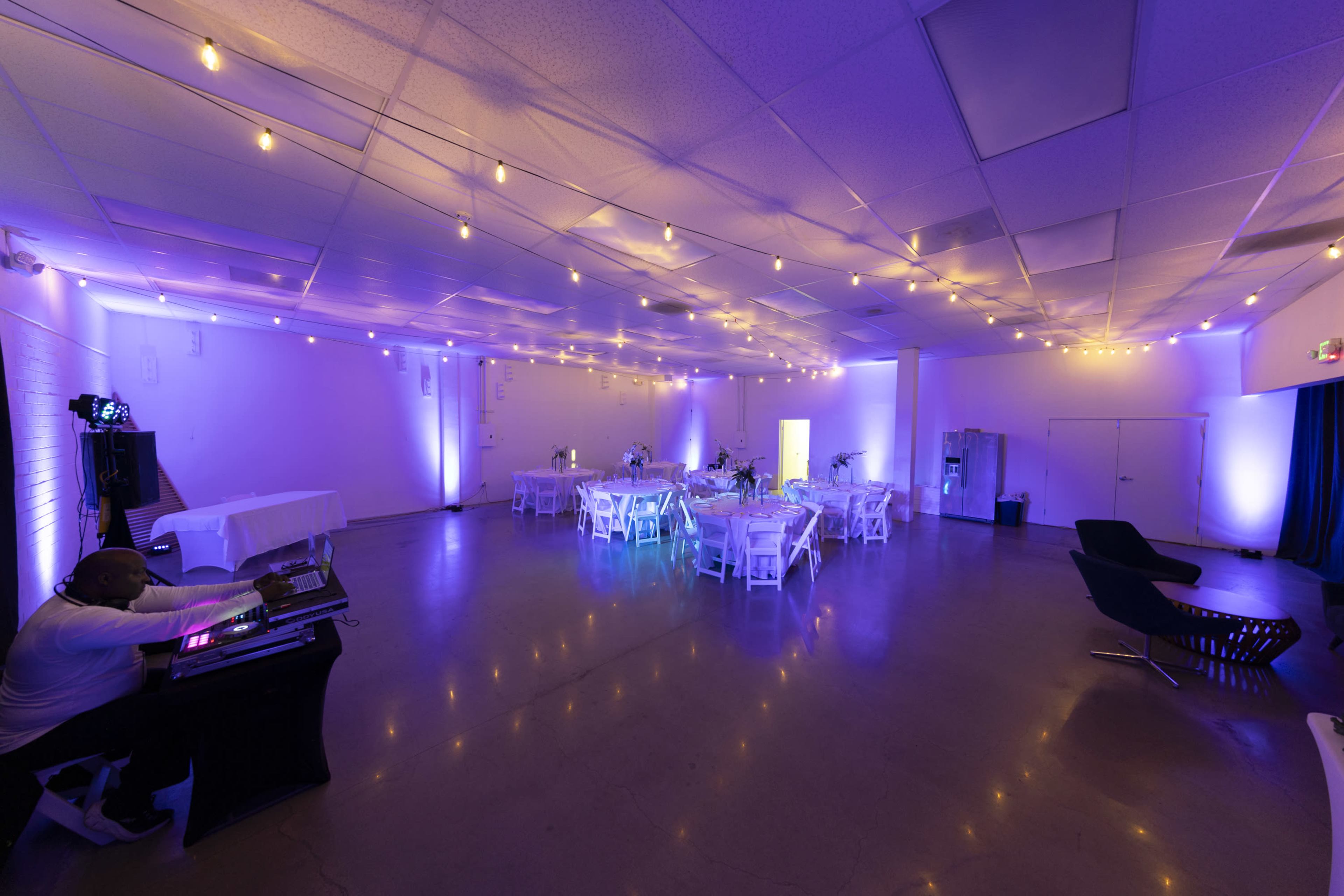 A spacious event venue features round tables with white tablecloths, decorated with centerpieces, and is illuminated by soft purple and warm white lighting, with string lights overhead.