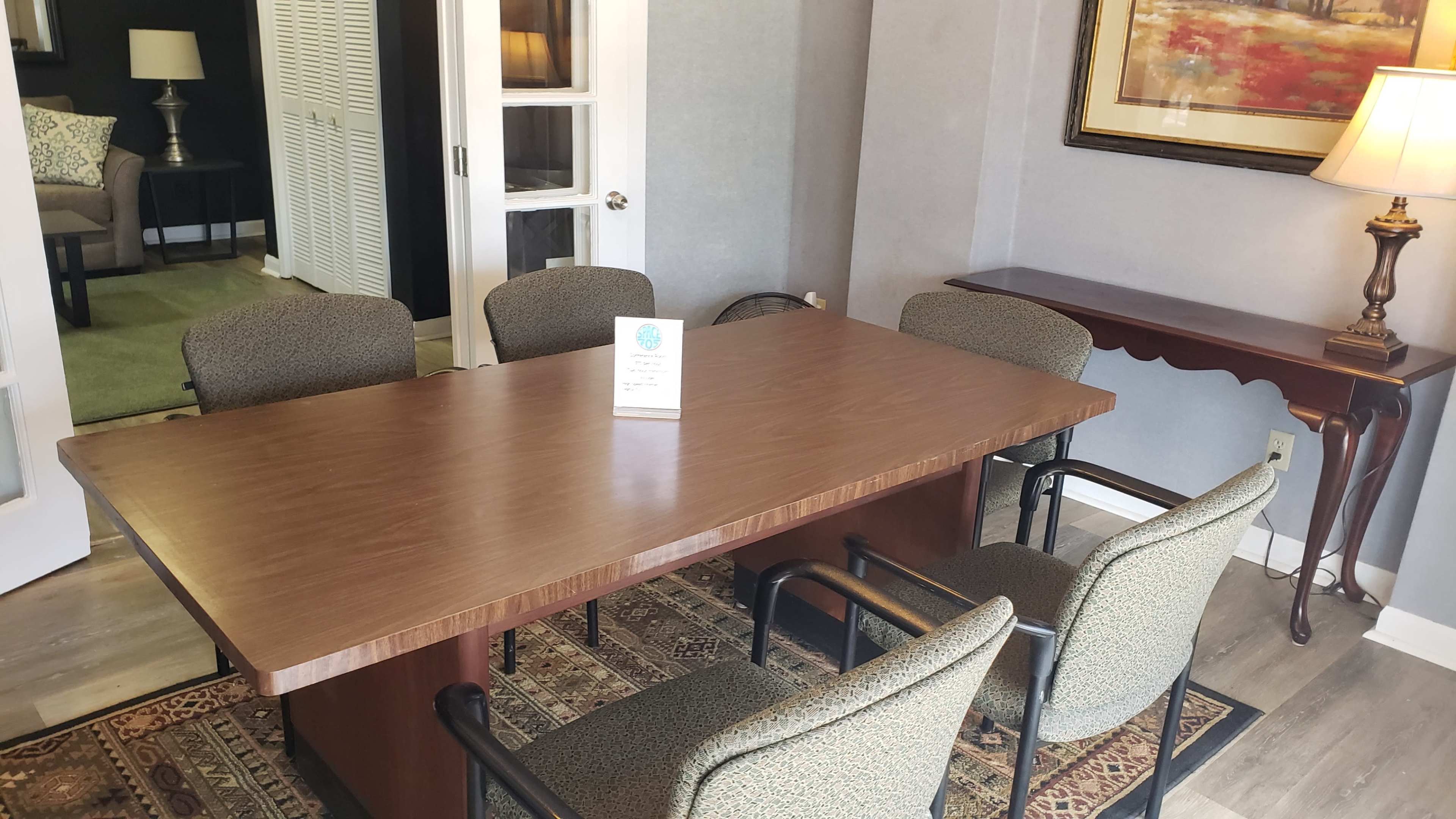 A rectangular wooden table surrounded by four upholstered chairs sits in a well-lit room, with a lamp and decorative artwork nearby.