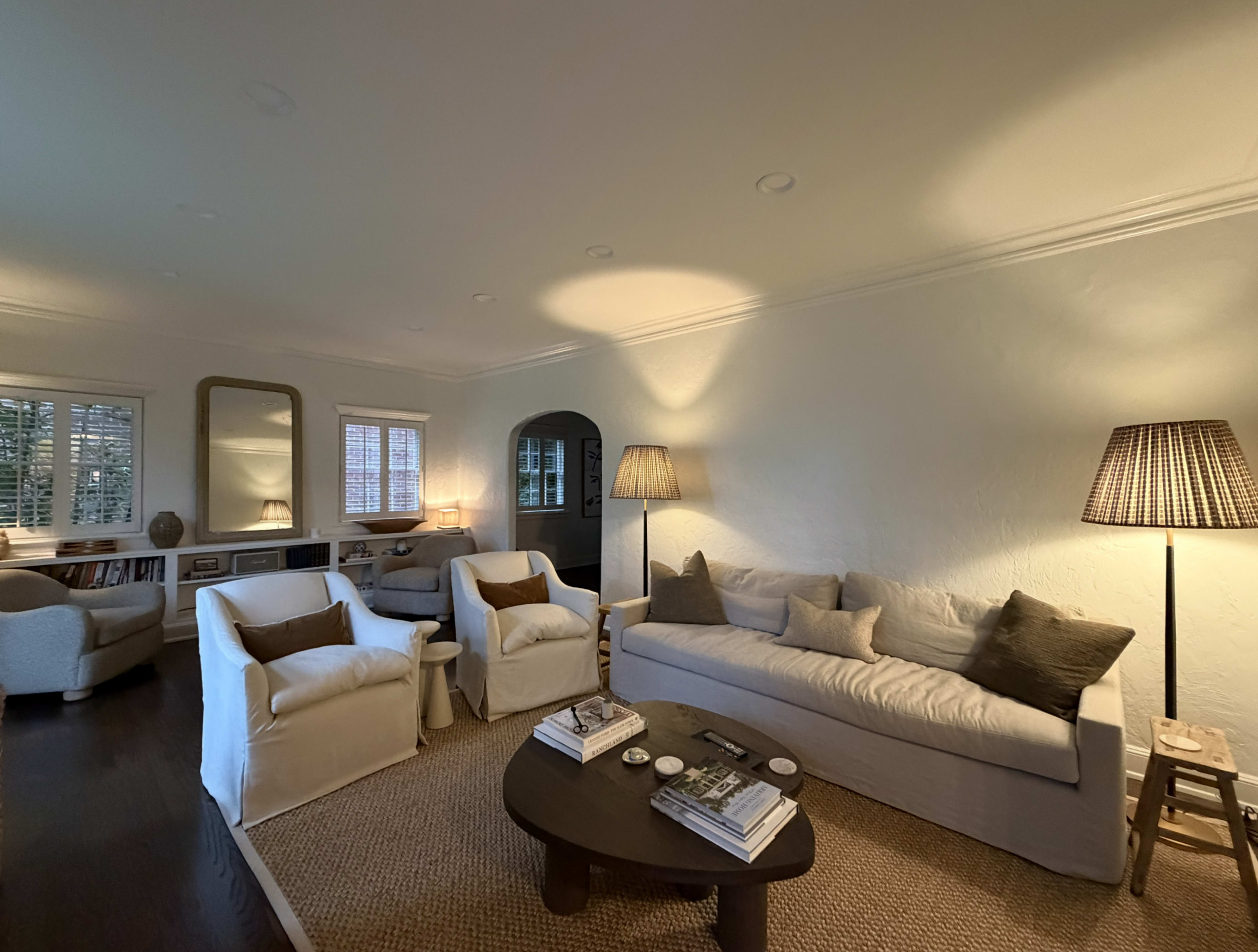 The image shows a cozy living room with a light-colored sofa, two armchairs, a coffee table, and lamps providing soft lighting.