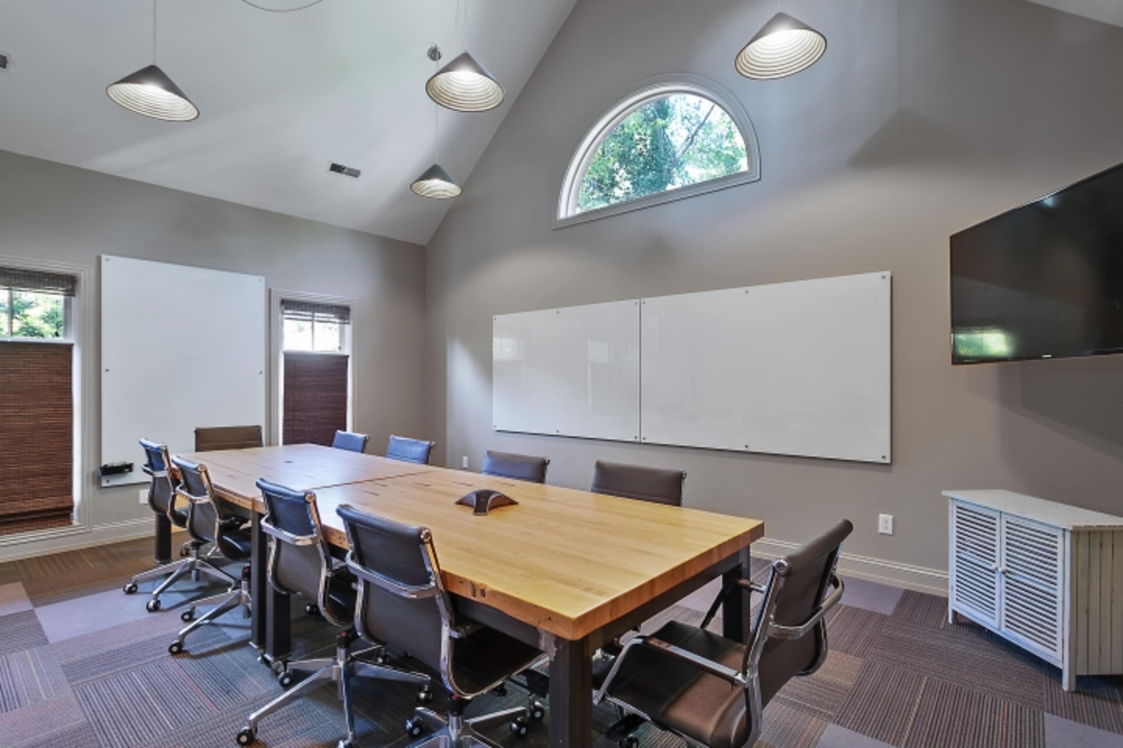 A conference room features a large wooden table surrounded by chairs, with whiteboards on the walls and windows letting in natural light.