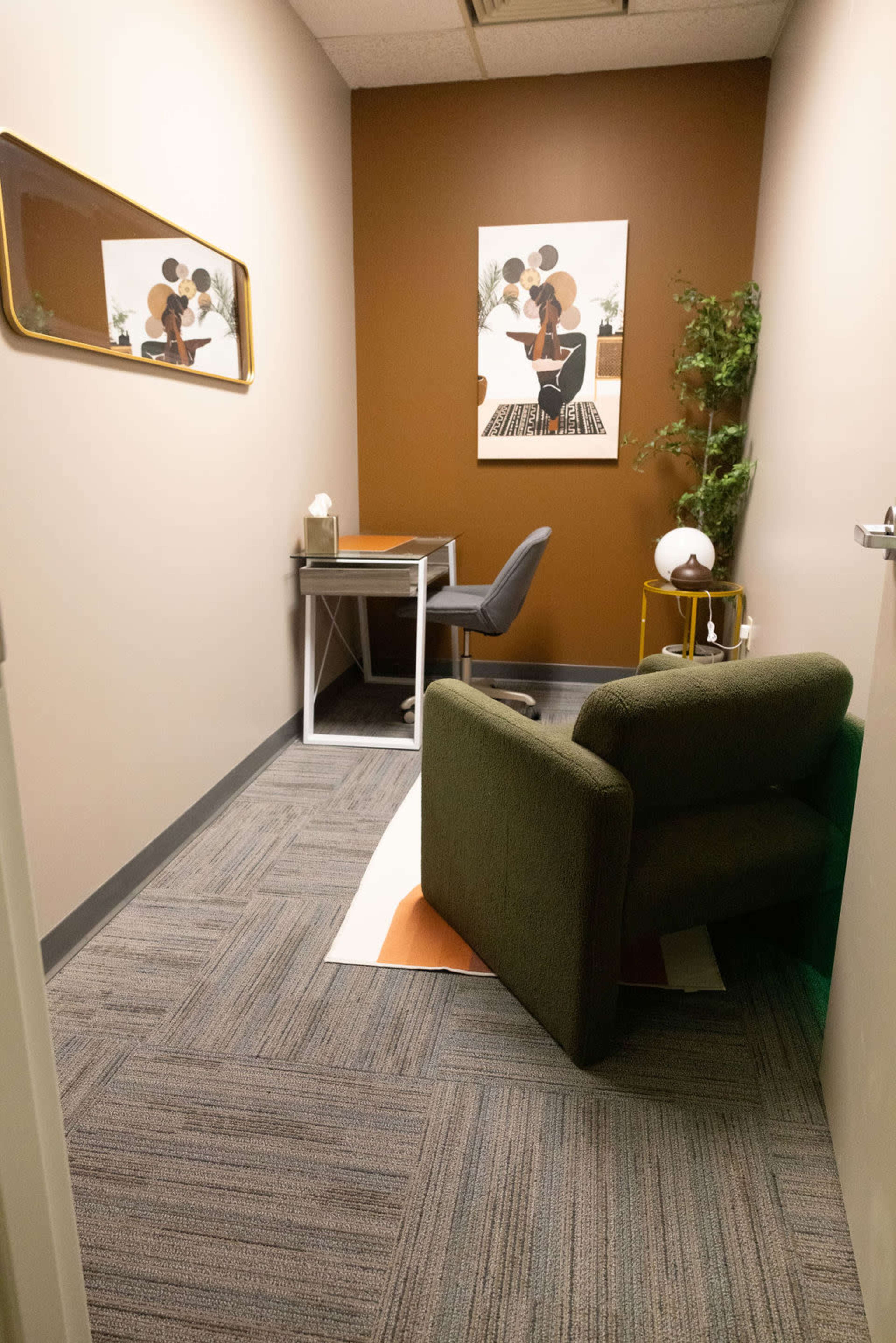 The image shows a small office space featuring a desk with a chair, a green armchair, a decorative plant, and a wall art piece, all set against a brown accent wall.