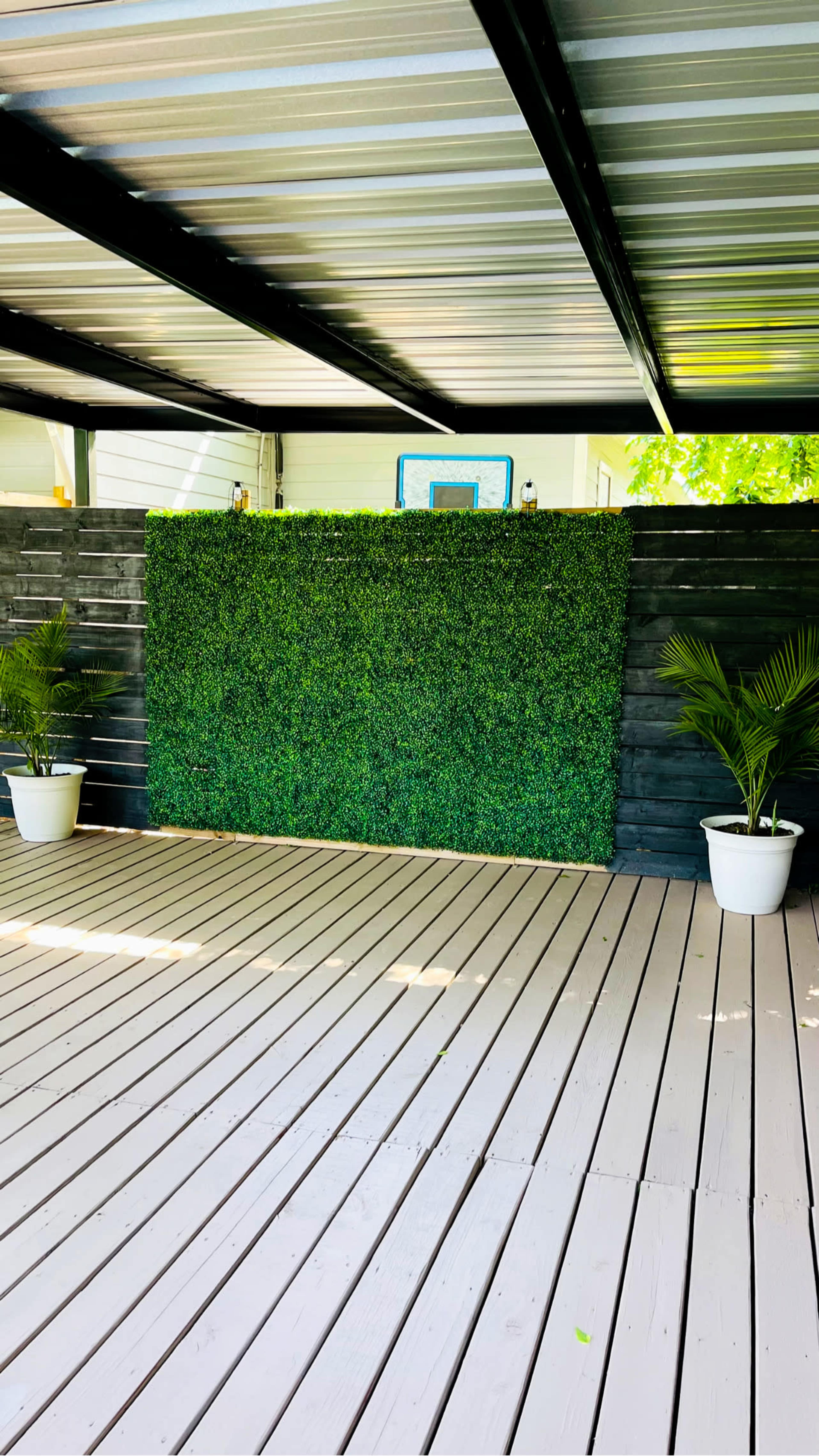 The image shows a wooden deck with a vertical faux hedge wall flanked by two white potted plants under a metal roof.