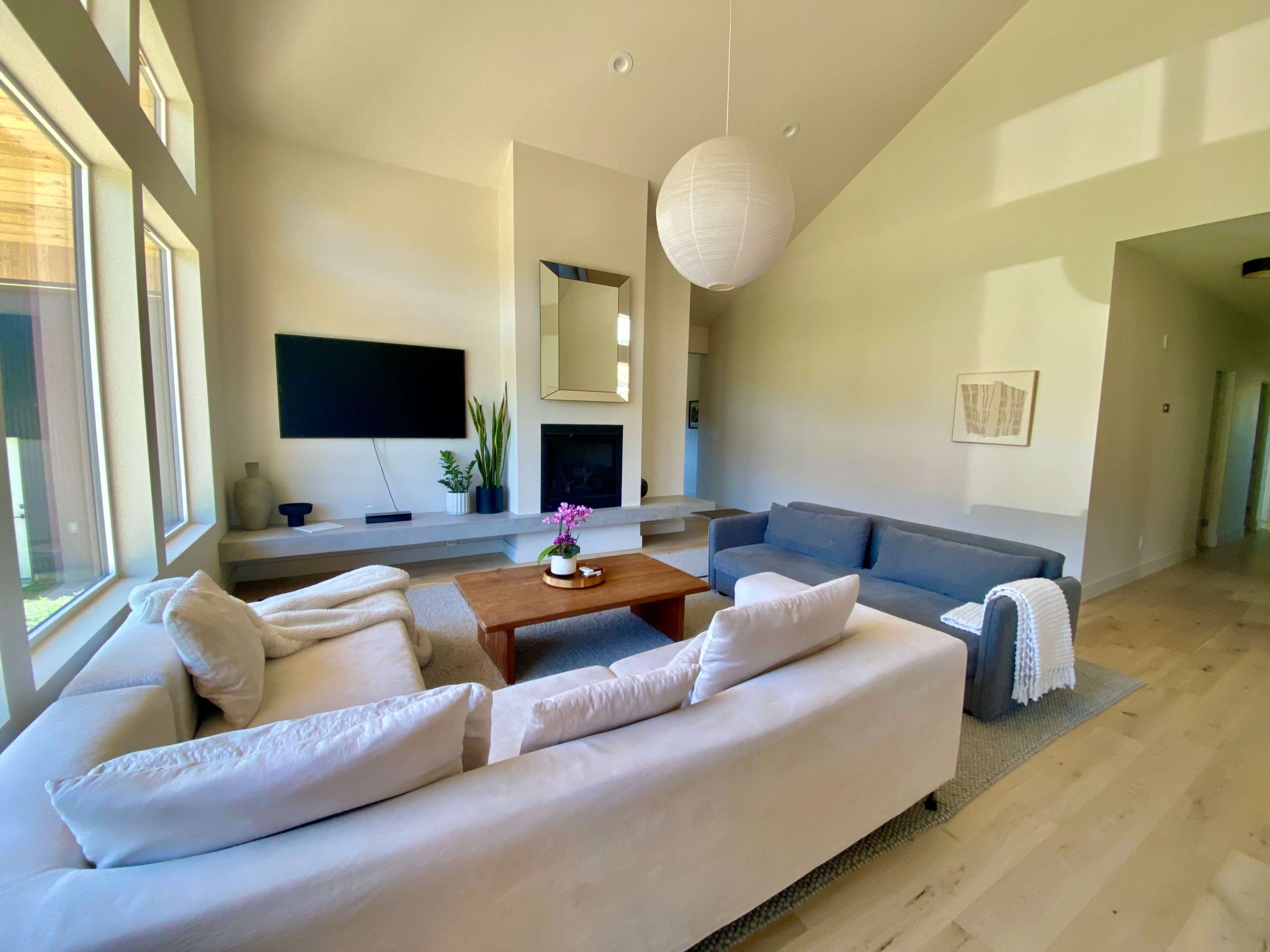 The image shows a bright living room with large windows, a television mounted on the wall, a coffee table with a vase of flowers, and two sofas positioned around the space.