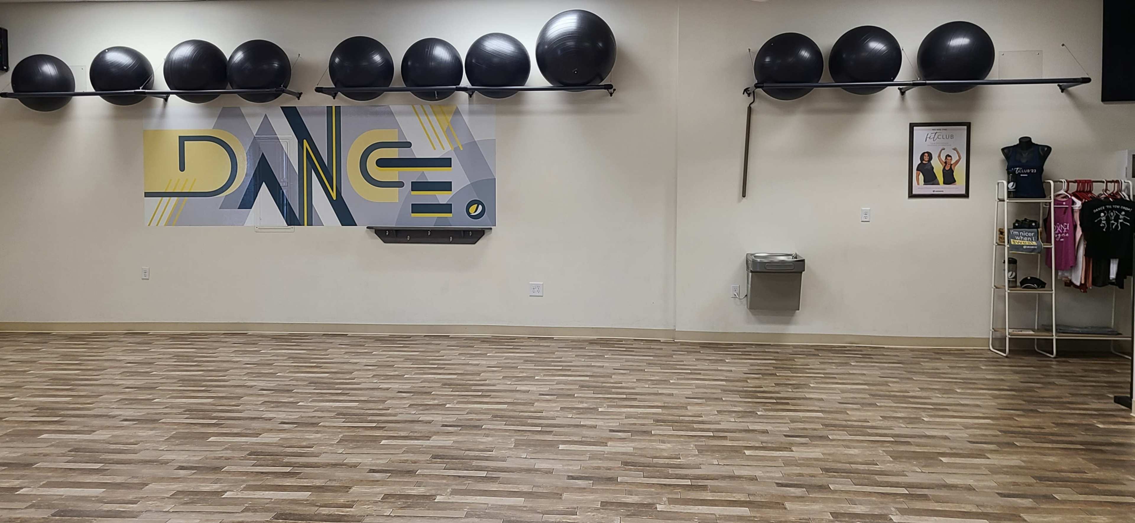 The image shows a dance studio with exercise balls on a rack, a water fountain, and a wall decorated with the word "DANCE."