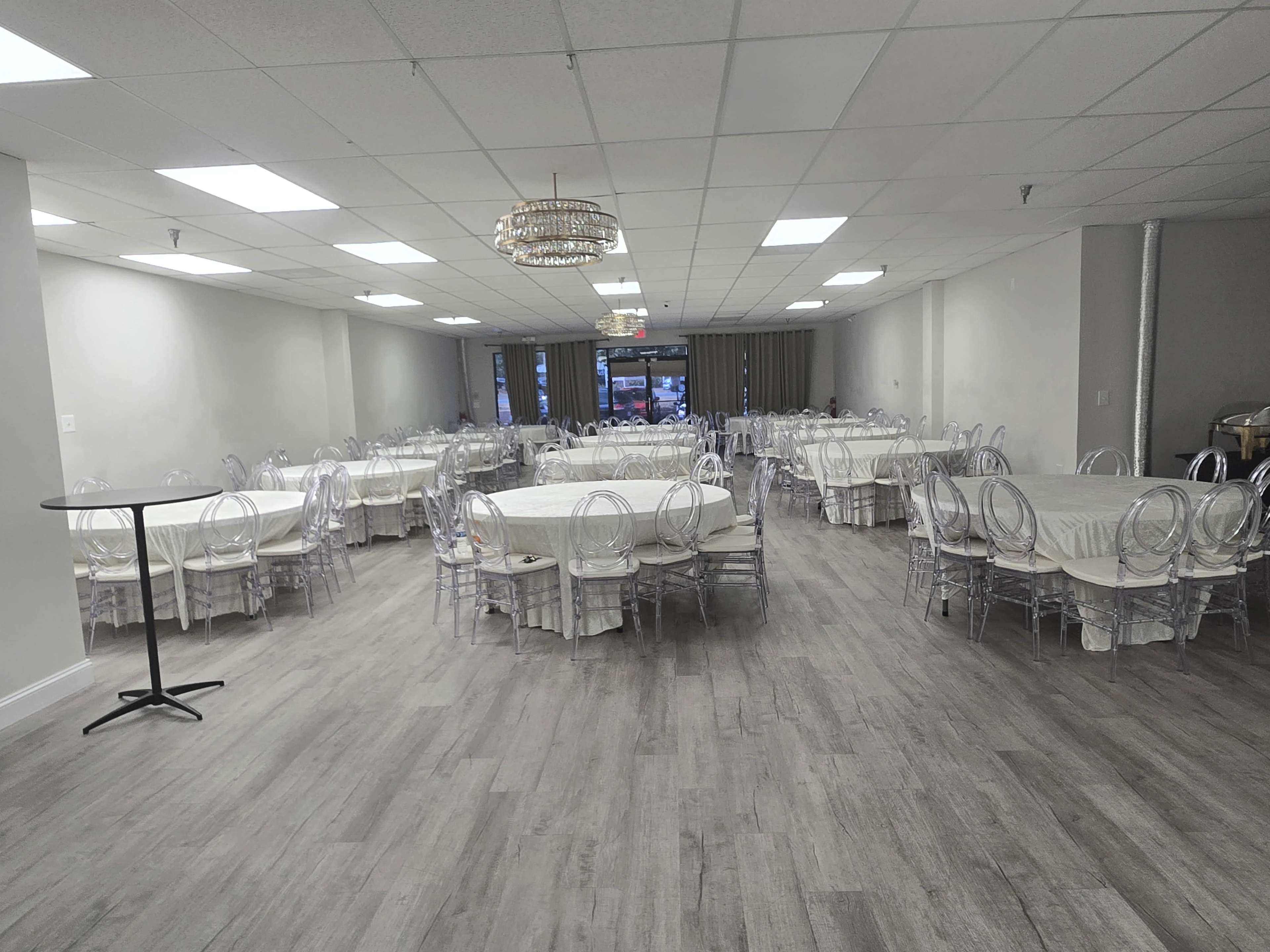 The image shows a large banquet hall with multiple round tables covered in white tablecloths and surrounded by clear ghost chairs.