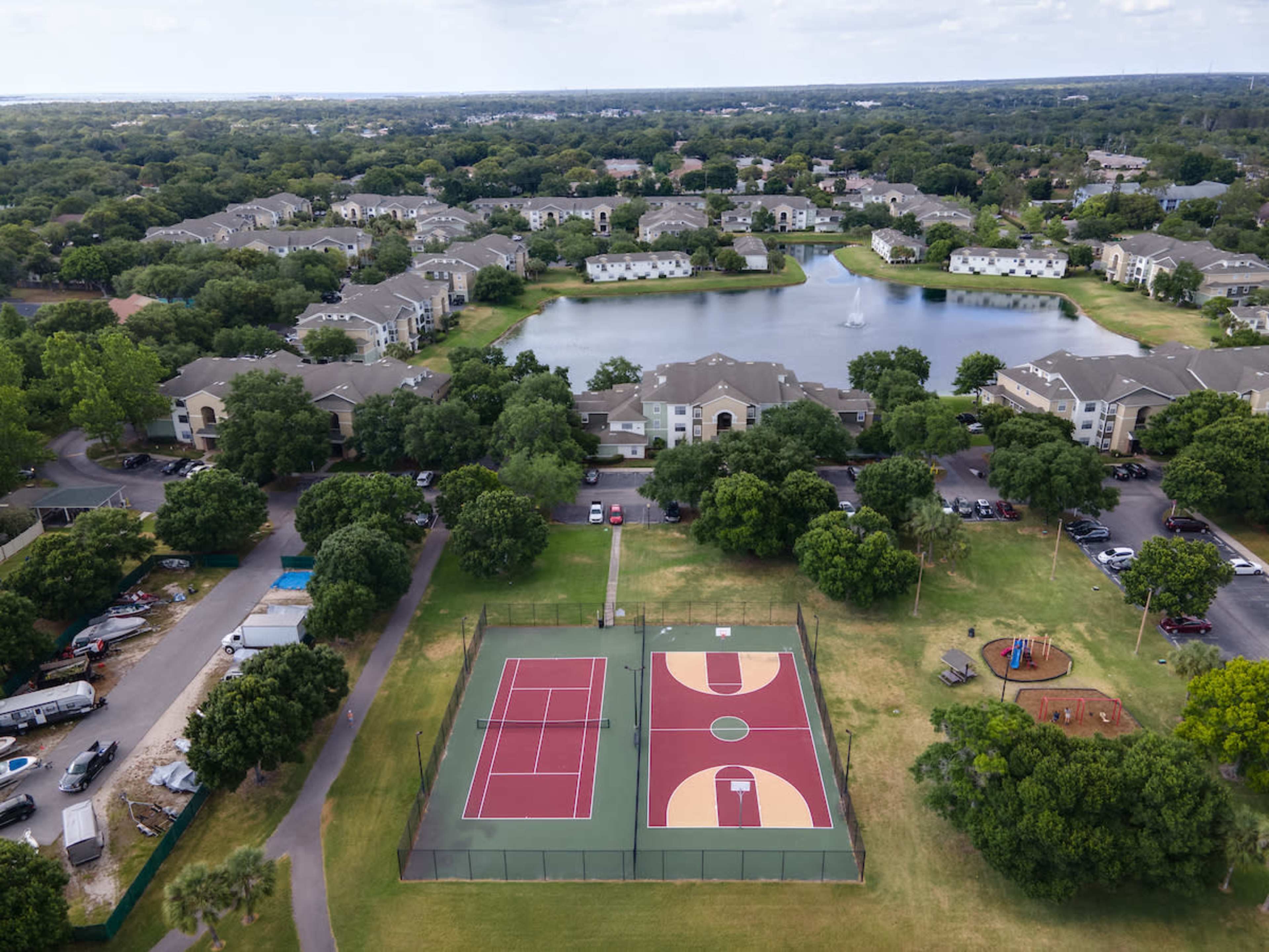 A tennis court surrounded by green lawns, trees, and residential apartment buildings near a pond.