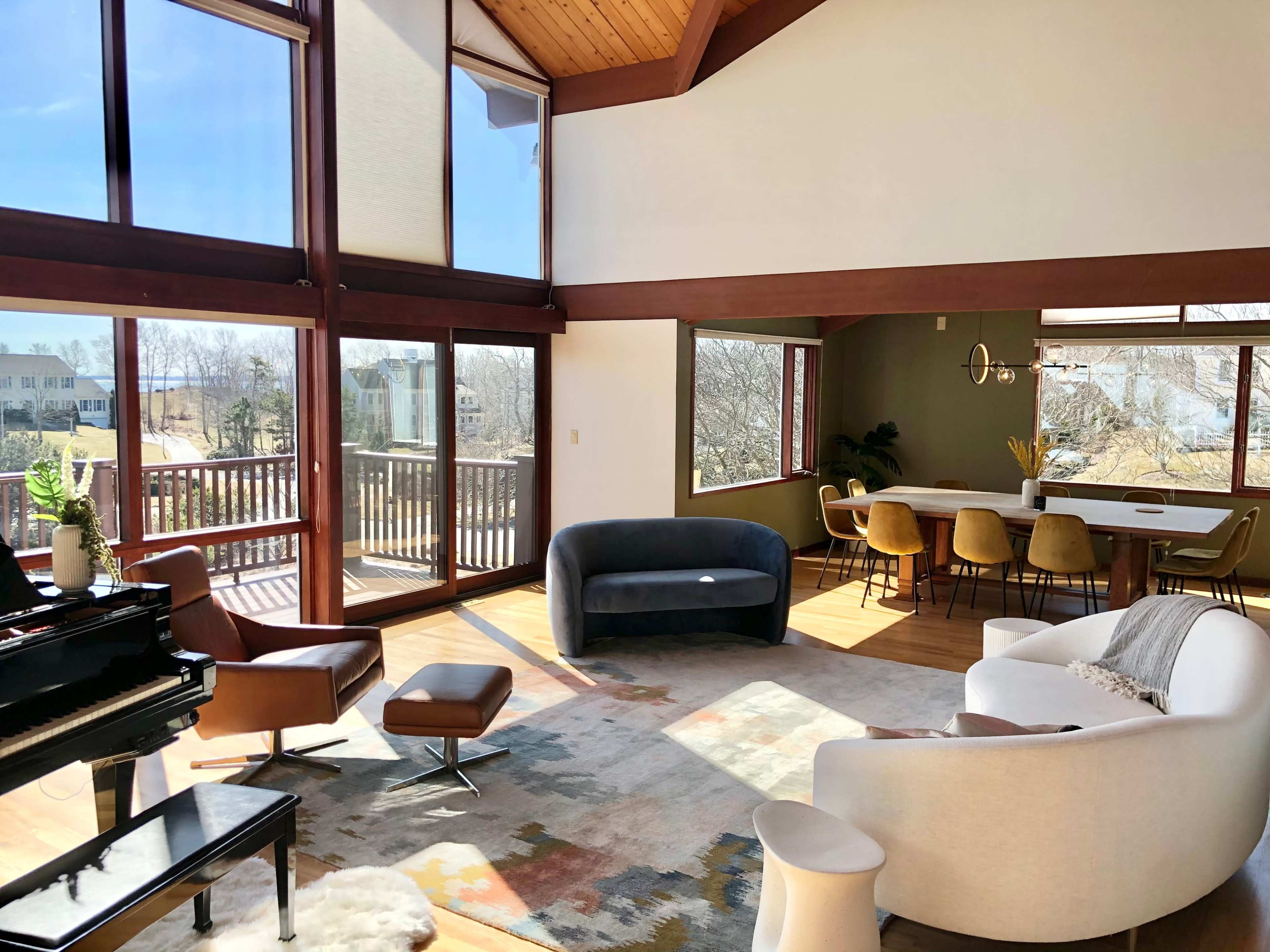 A modern living space featuring large windows, a piano, and various seating options, with a dining area visible in the background.