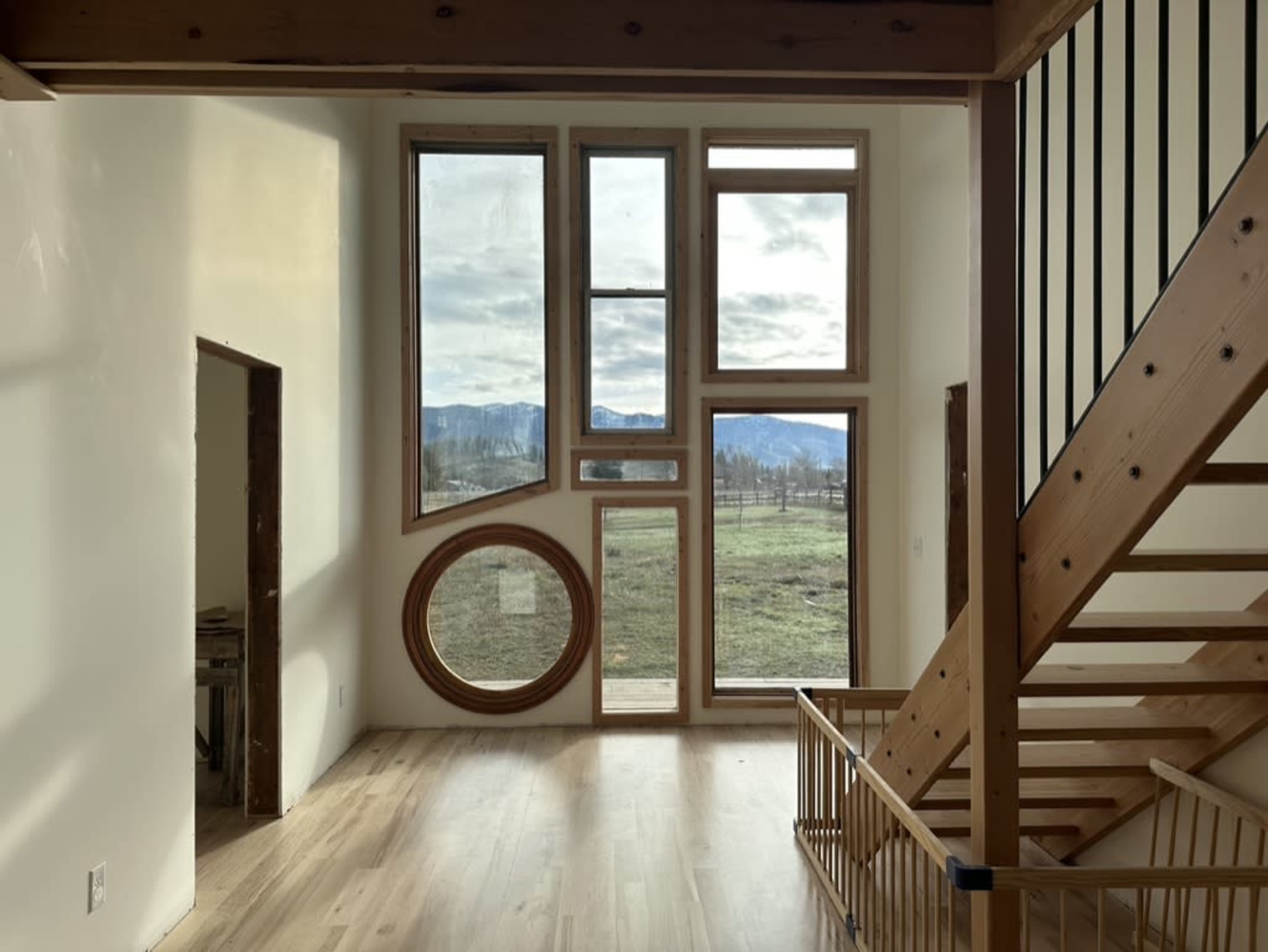 The image shows a room with multiple large windows, including a circular one, and a wooden staircase leading to an upper level.