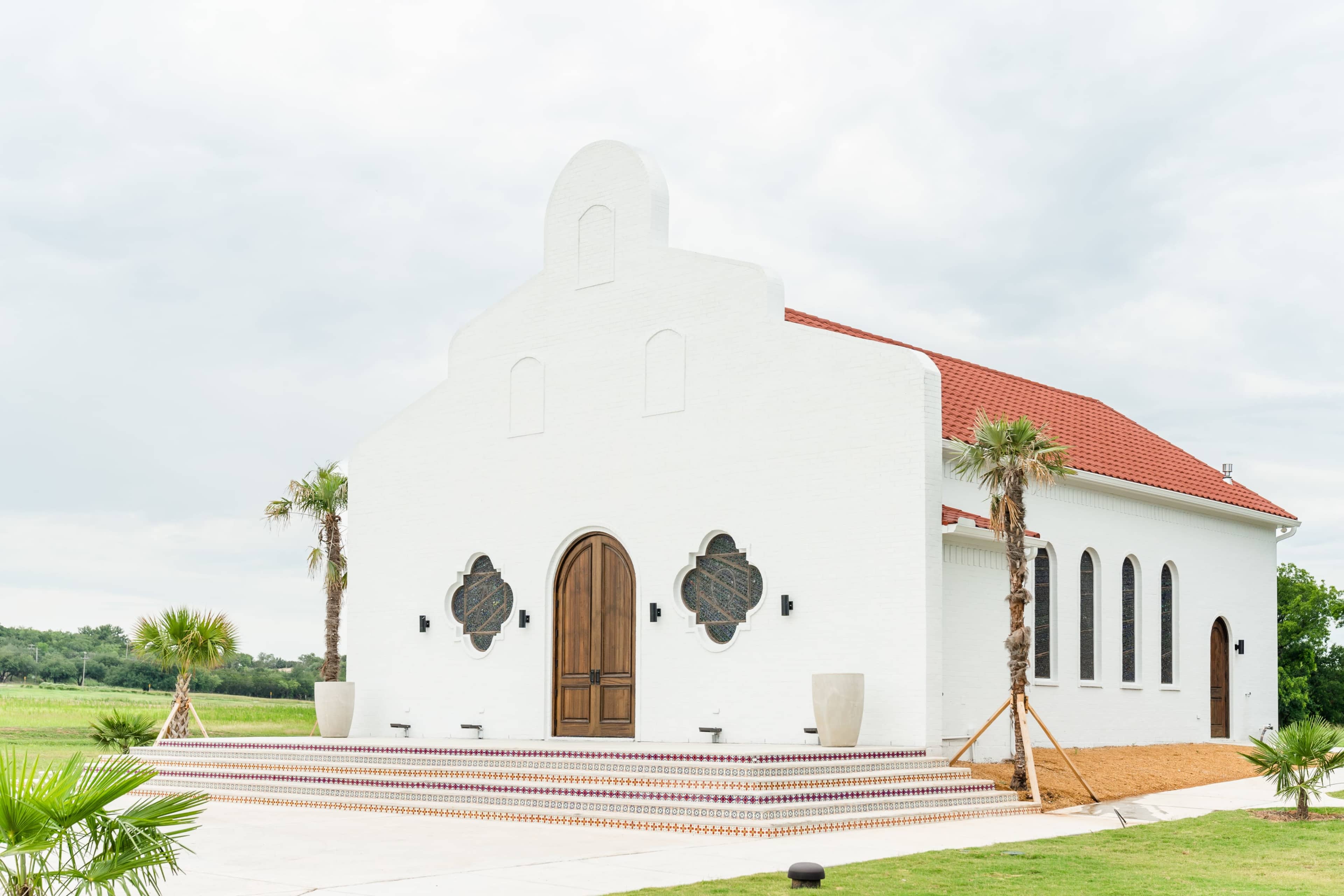 A white, structured building with a red roof features large arched windows and is surrounded by landscaped greenery.