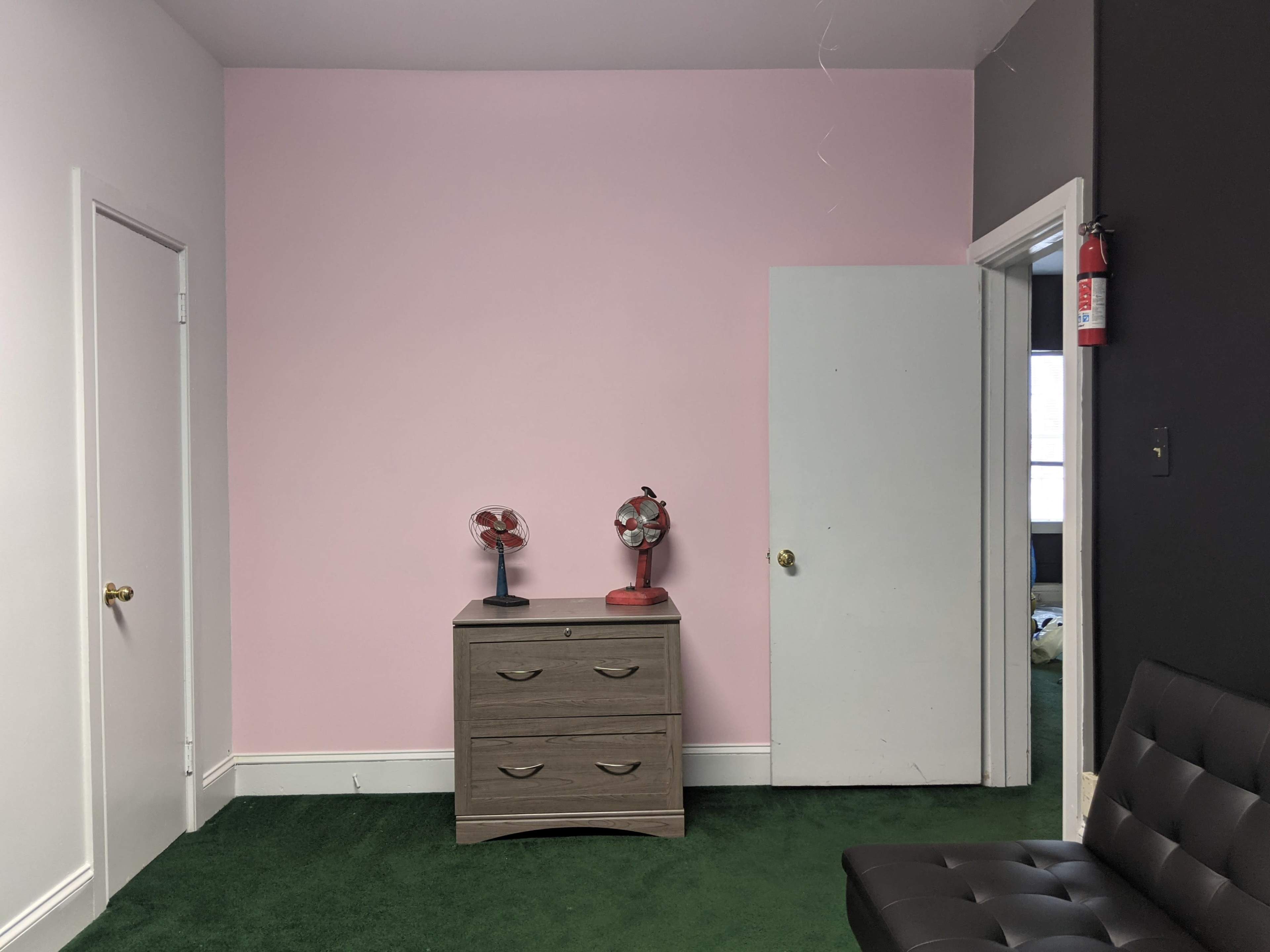 A room with a pink wall, a gray dresser with drawers, two small fans on top of the dresser, a green carpet, and a partially open white door.