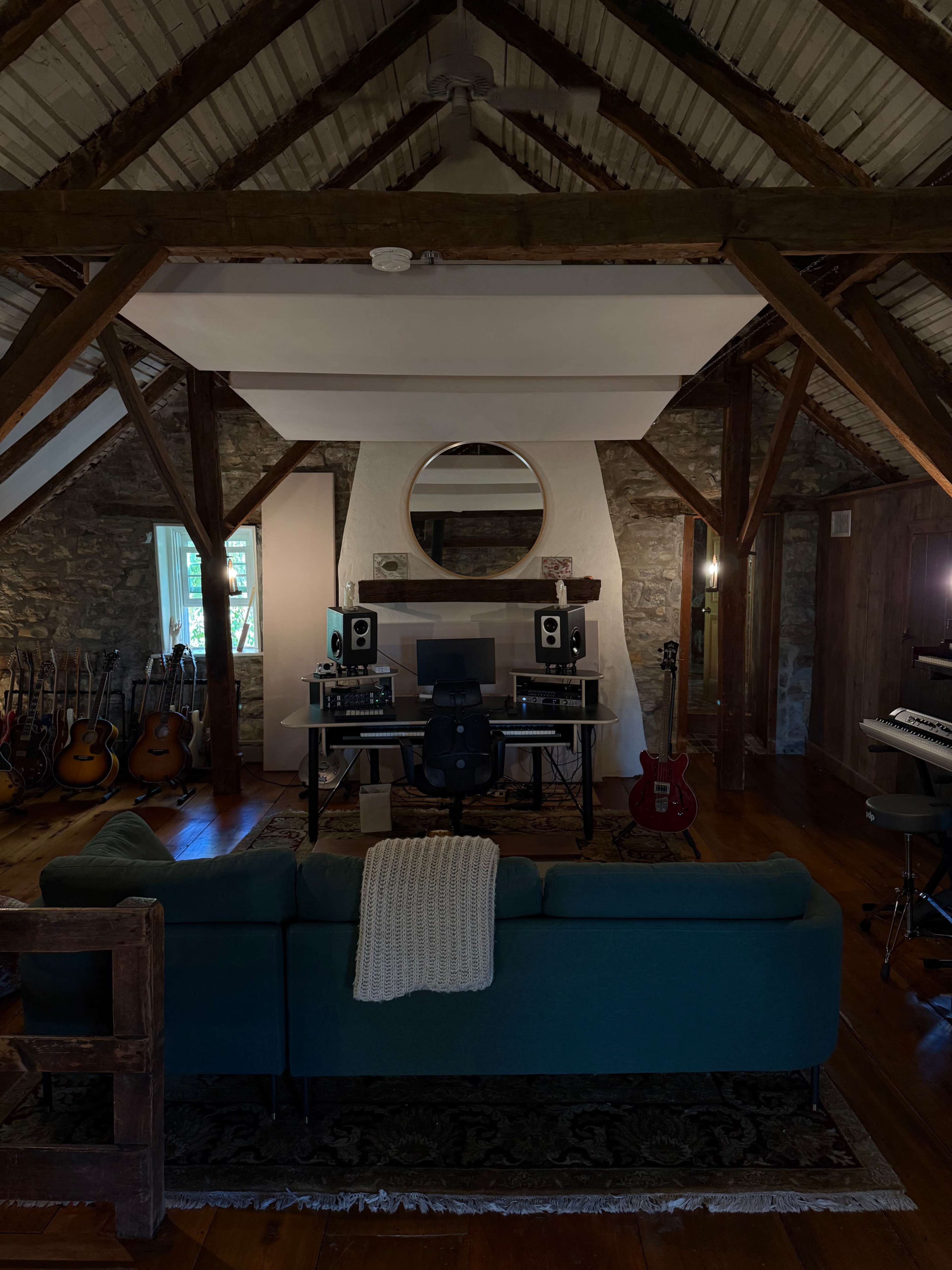 The image shows a cozy music studio with a modern setup, featuring a desk with audio equipment, surrounded by wooden beams and stone walls, and musical instruments displayed in the background.