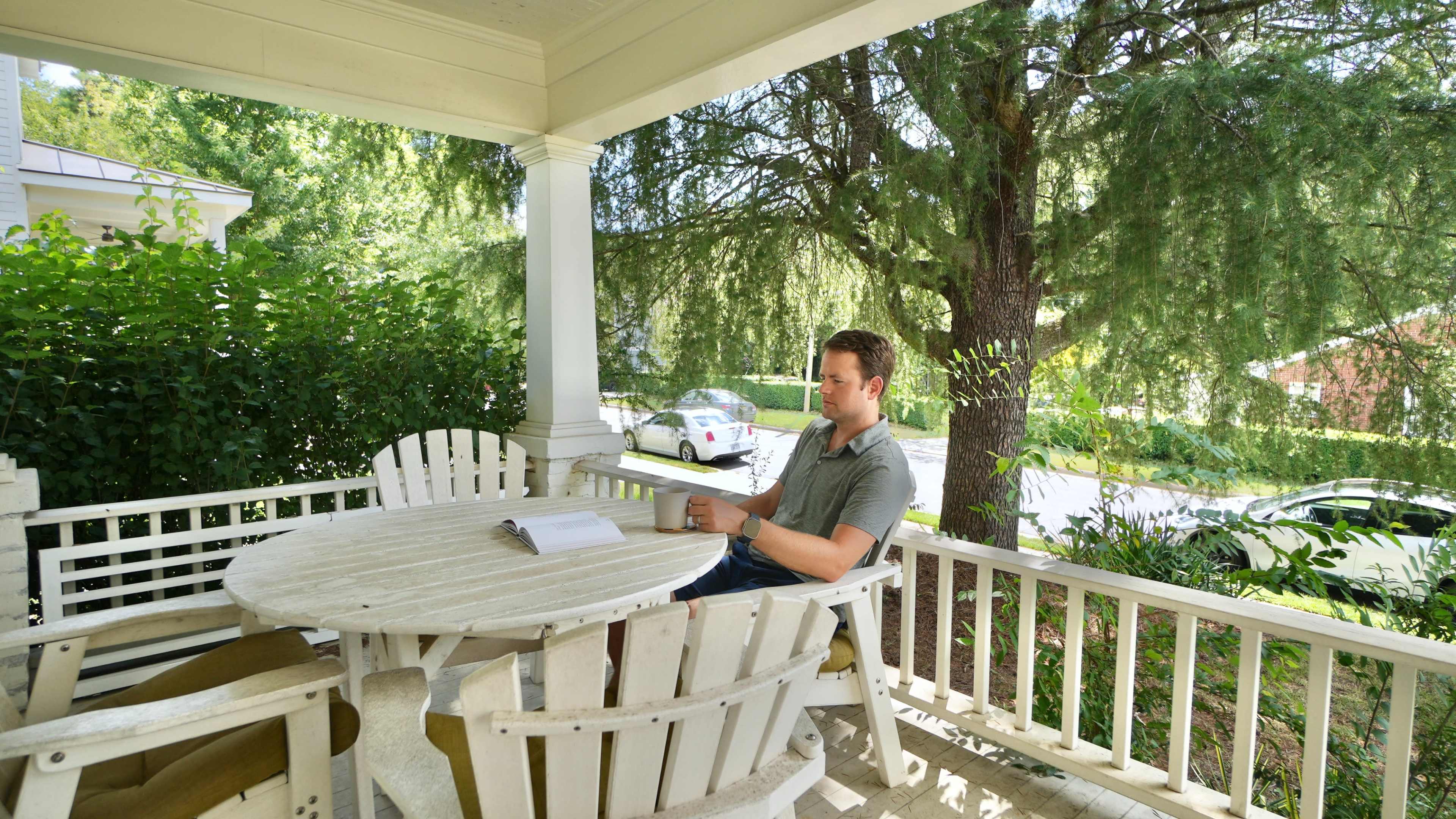 A man sits at a white wooden table on a porch, reading a book while surrounded by green foliage.