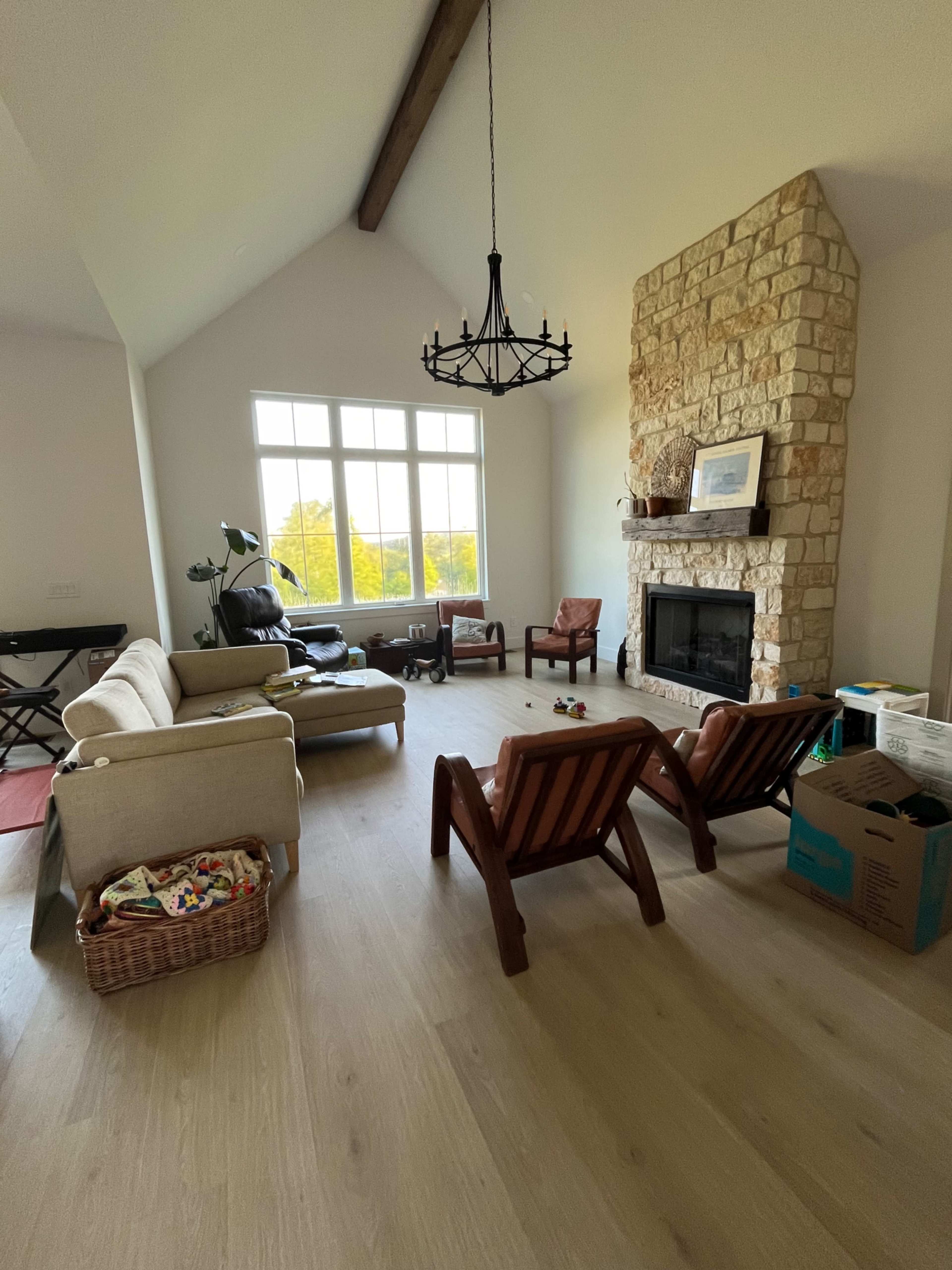 The image shows a spacious living room featuring a stone fireplace, large windows, a chandelier, and wooden chairs arranged around a light-colored sofa.