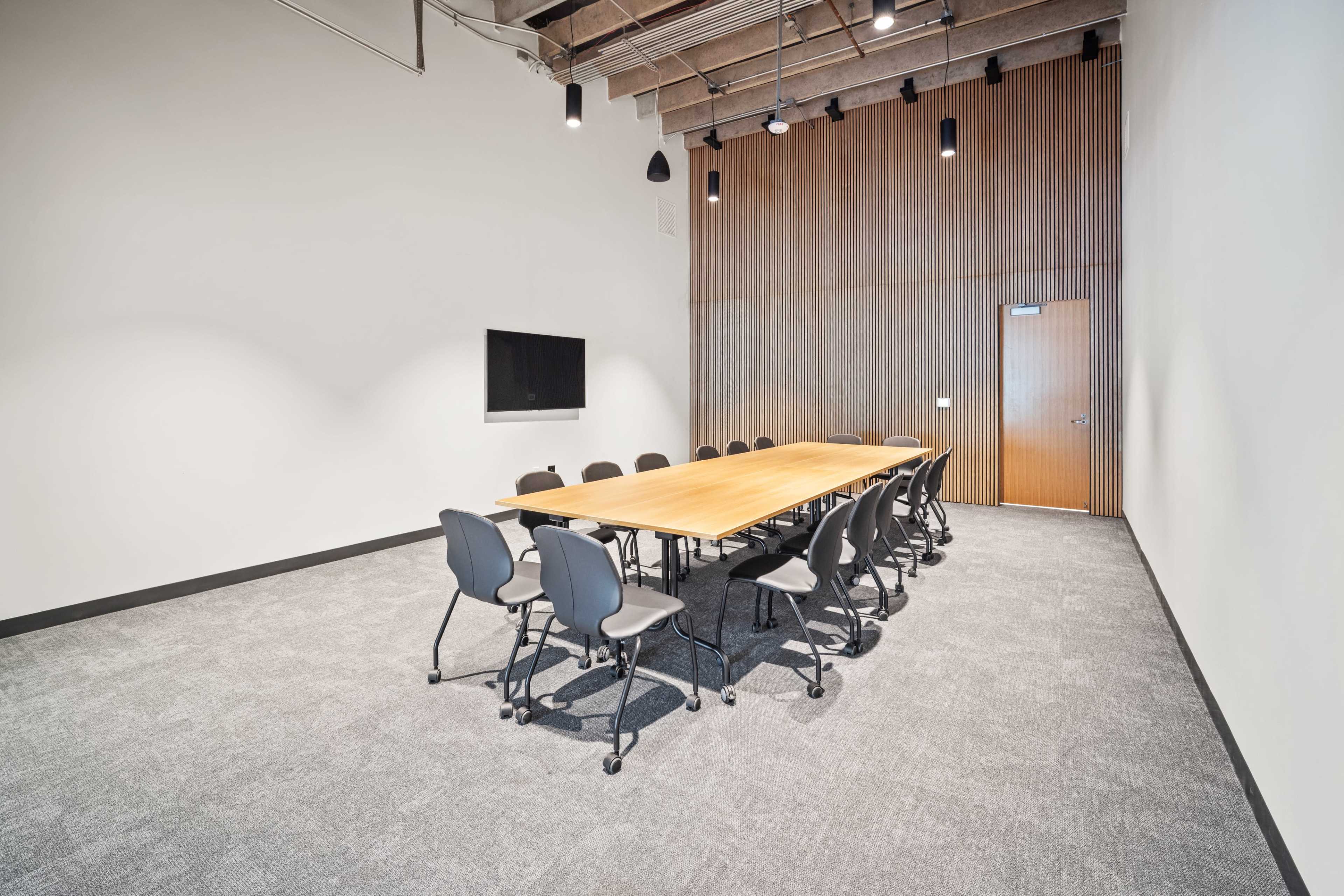 The image shows a modern conference room featuring a long wooden table surrounded by several office chairs, with a wall-mounted television and a wooden accent wall.