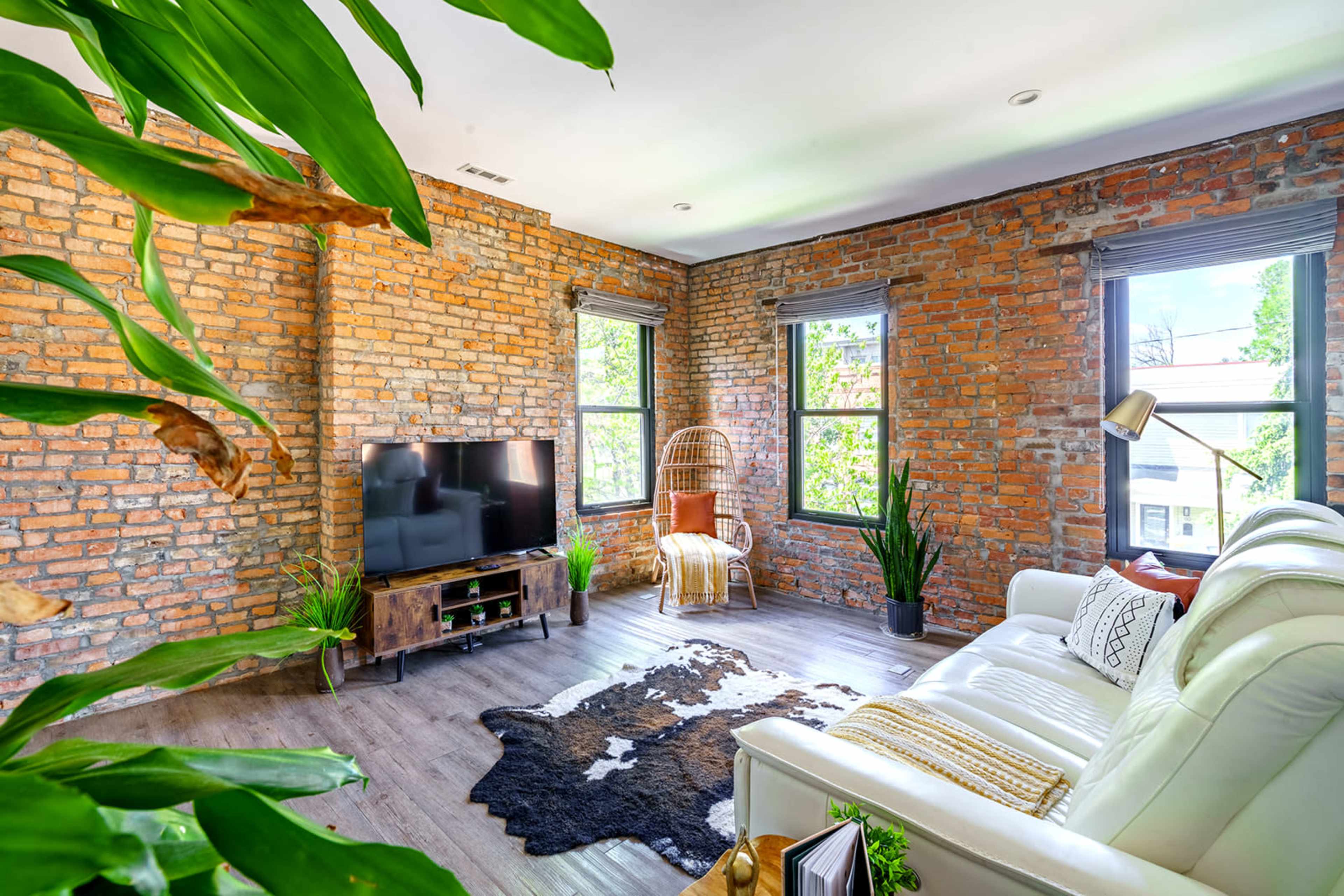A modern living room features a white sofa, a wooden TV stand, and large windows that illuminate exposed brick walls and greenery.