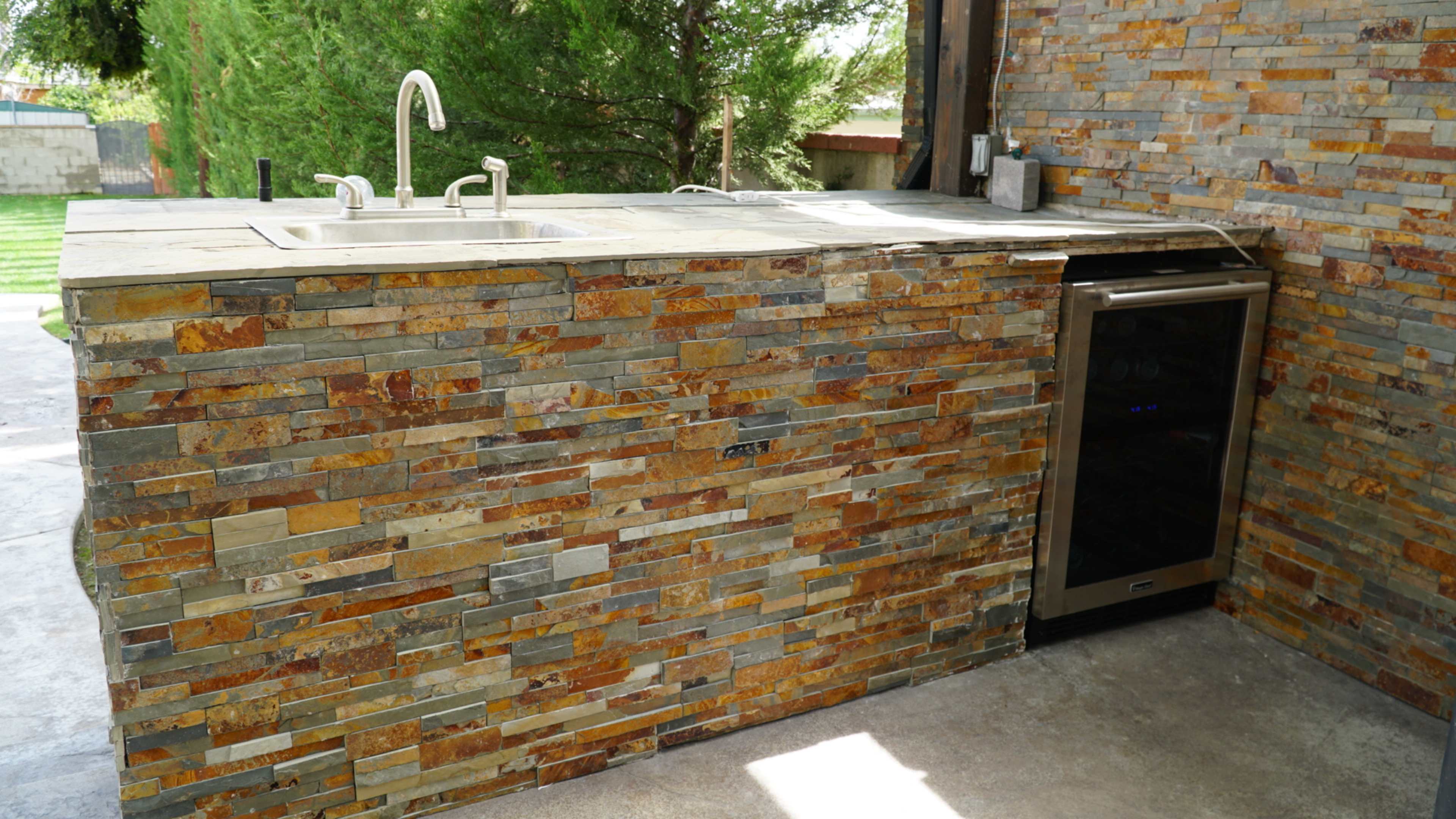 The image shows an outdoor kitchen area with a stone countertop, a sink, and a refrigerator built into the cabinetry.