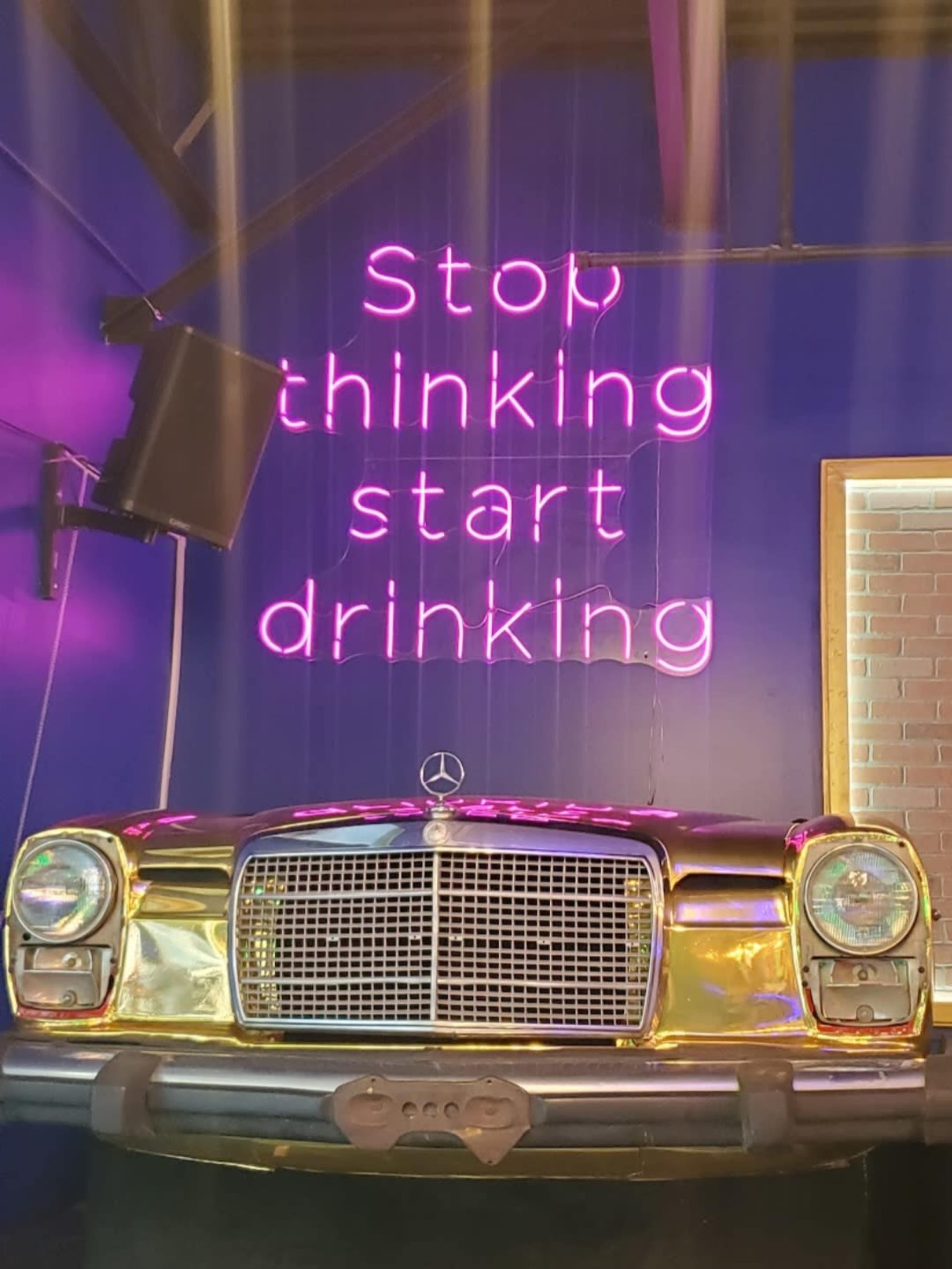 A vintage Mercedes-Benz car sits under a neon sign that reads, "Stop thinking start drinking," against a blue wall.