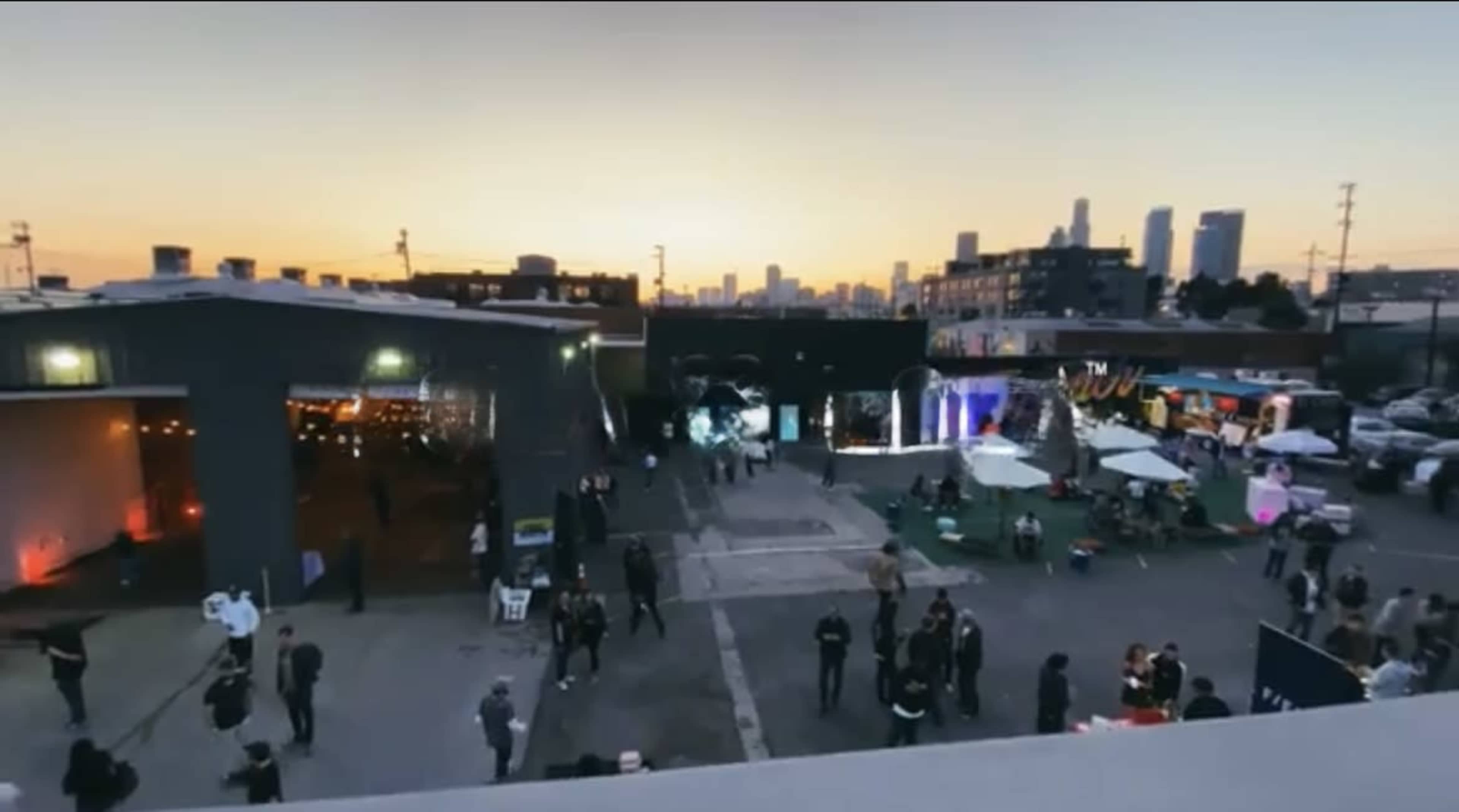 The image shows an outdoor event space at dusk with people gathered, food stalls, and city skyline in the background.