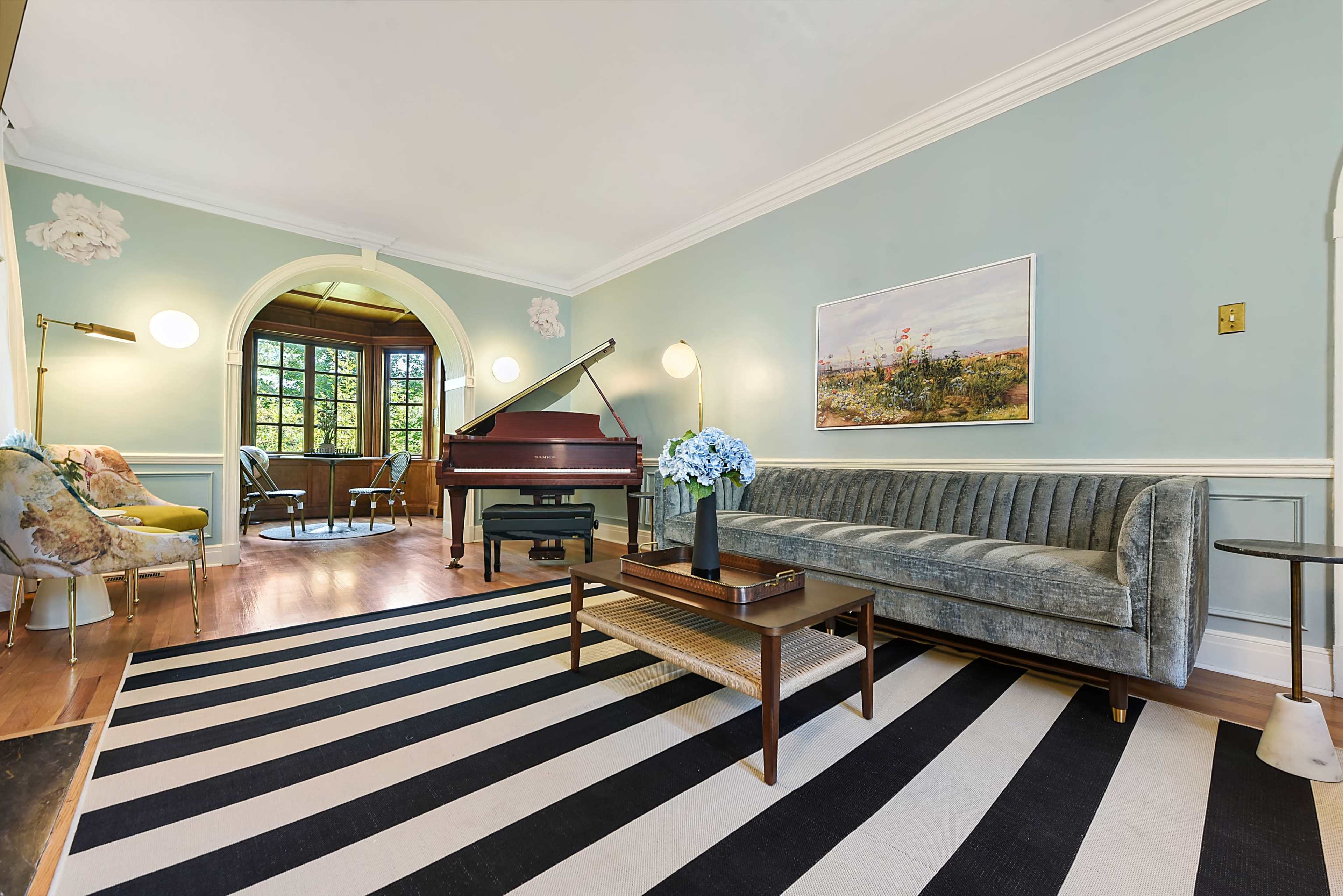 The image depicts a brightly lit living room featuring a grand piano, a striped area rug, a modern sofa, and a small coffee table adorned with flowers.