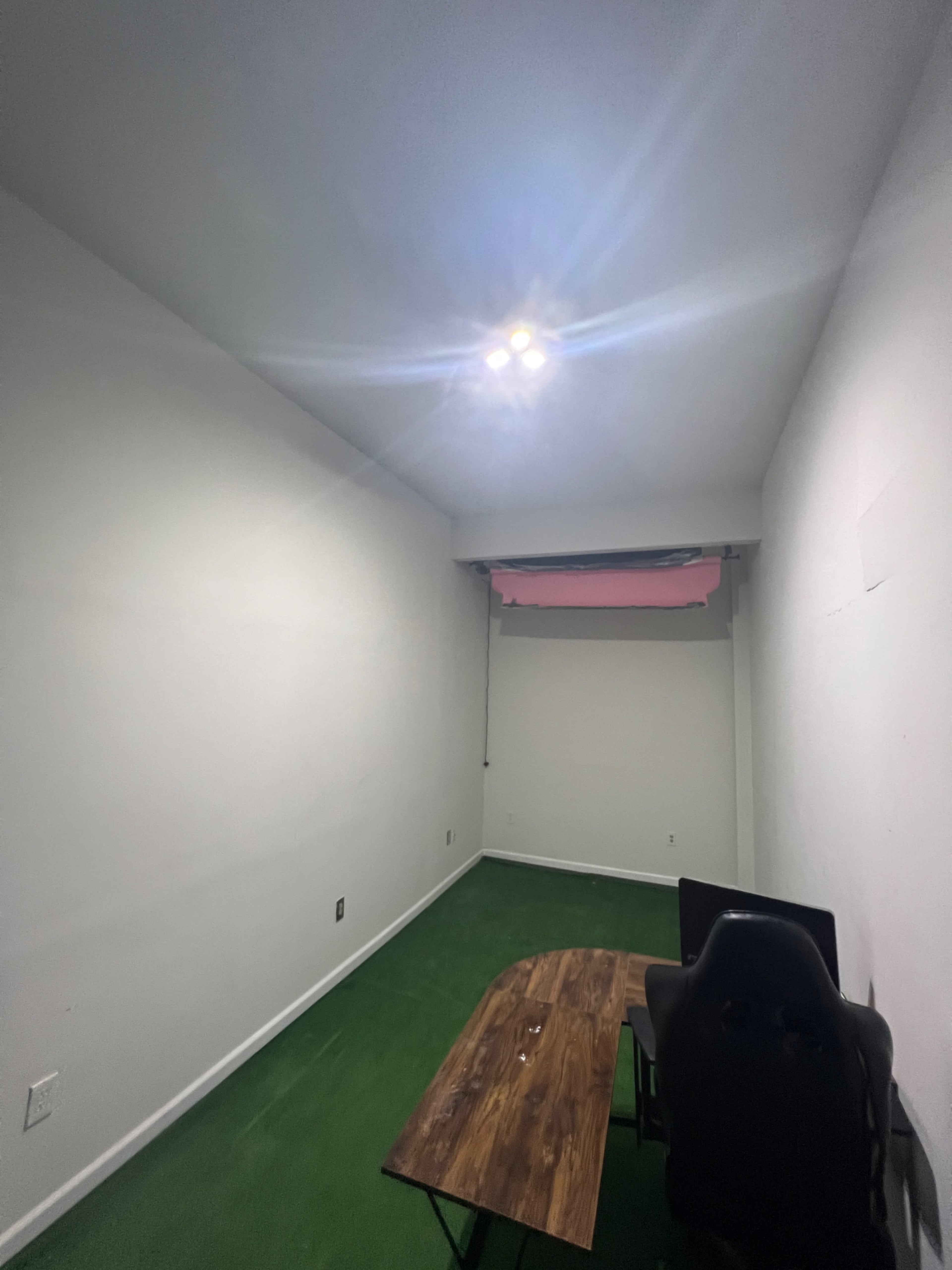 A sparsely furnished room with a wooden table and a black chair on a green carpet, illuminated by recessed lights.