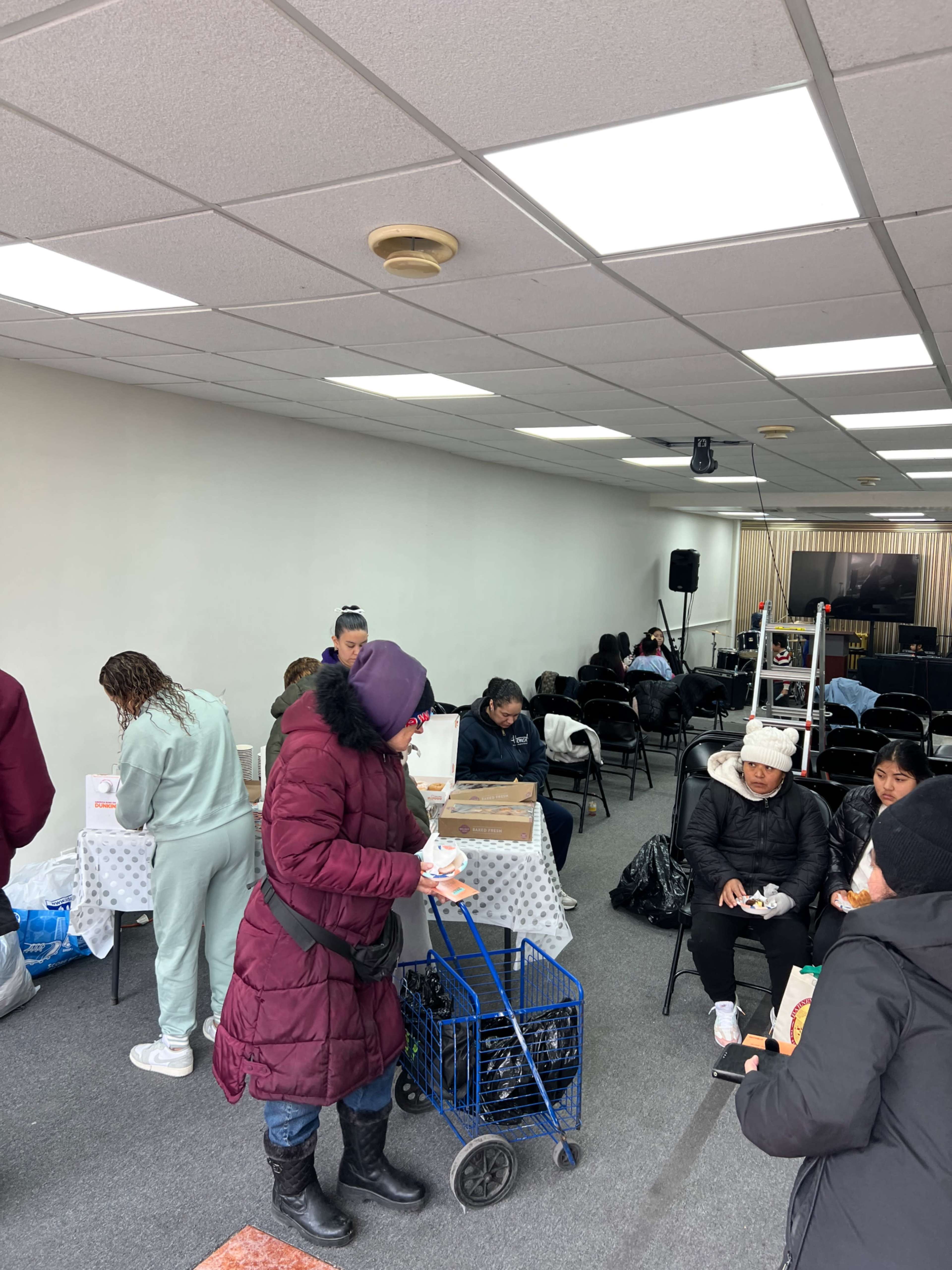 The scene shows a group of people inside a room with chairs and tables, where some individuals are sitting while others are standing and interacting, and a cart is parked nearby.