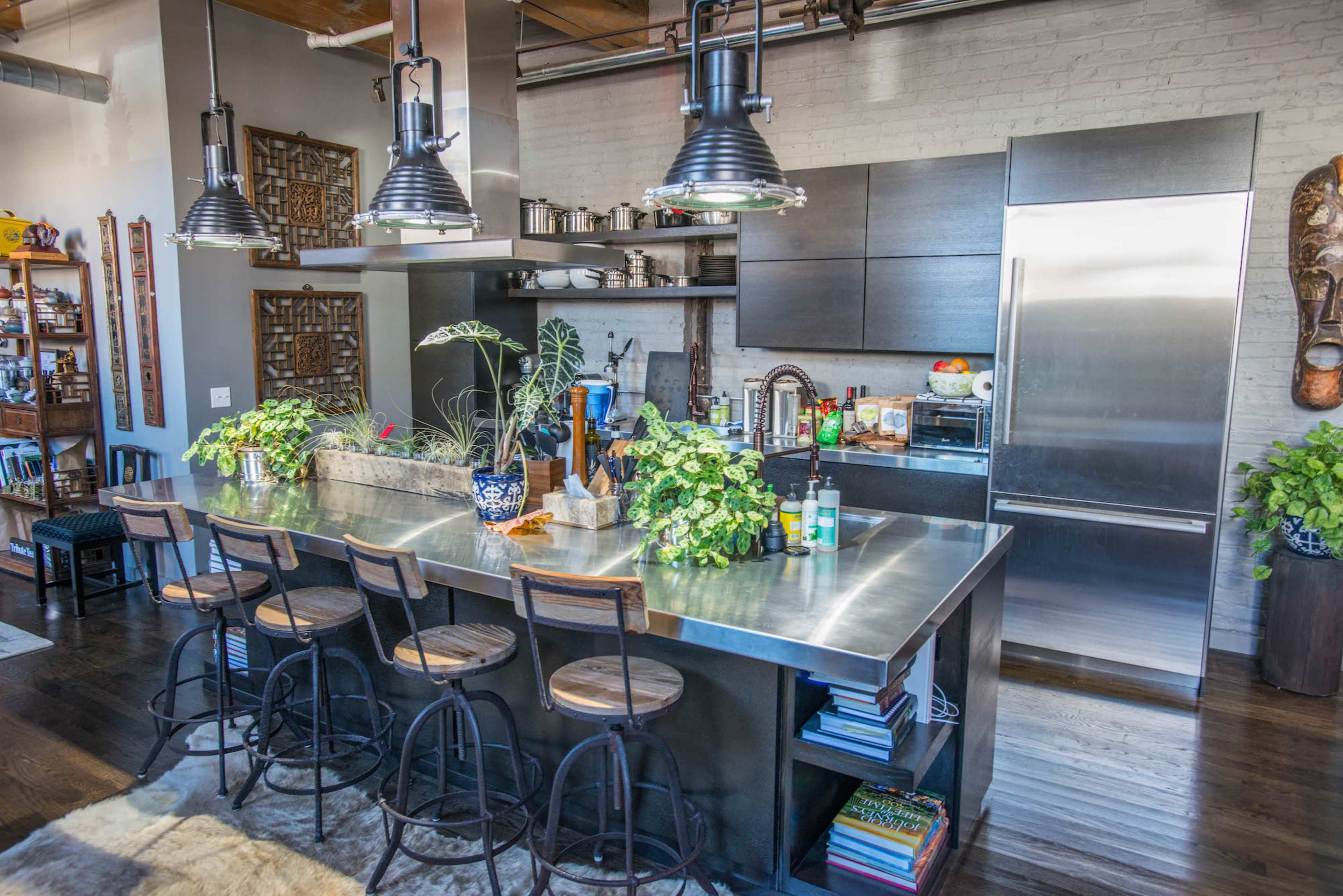 A modern kitchen with a central metal island, bar stools, and various plants, set against a backdrop of dark cabinetry and stainless steel appliances.