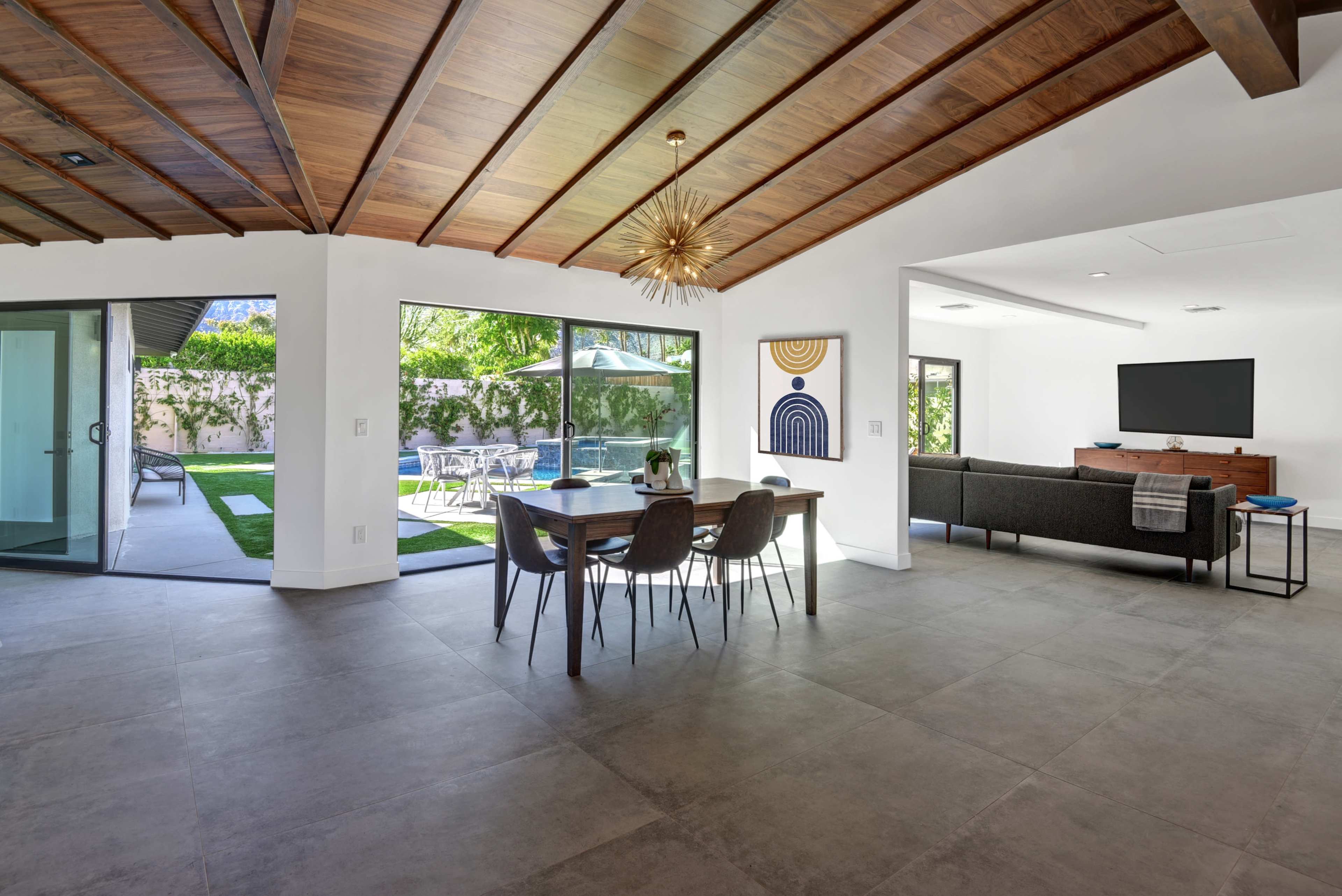 An open-concept living space with a dining area featuring a round table and black chairs, leading to a backyard with patio furniture and a pool area.