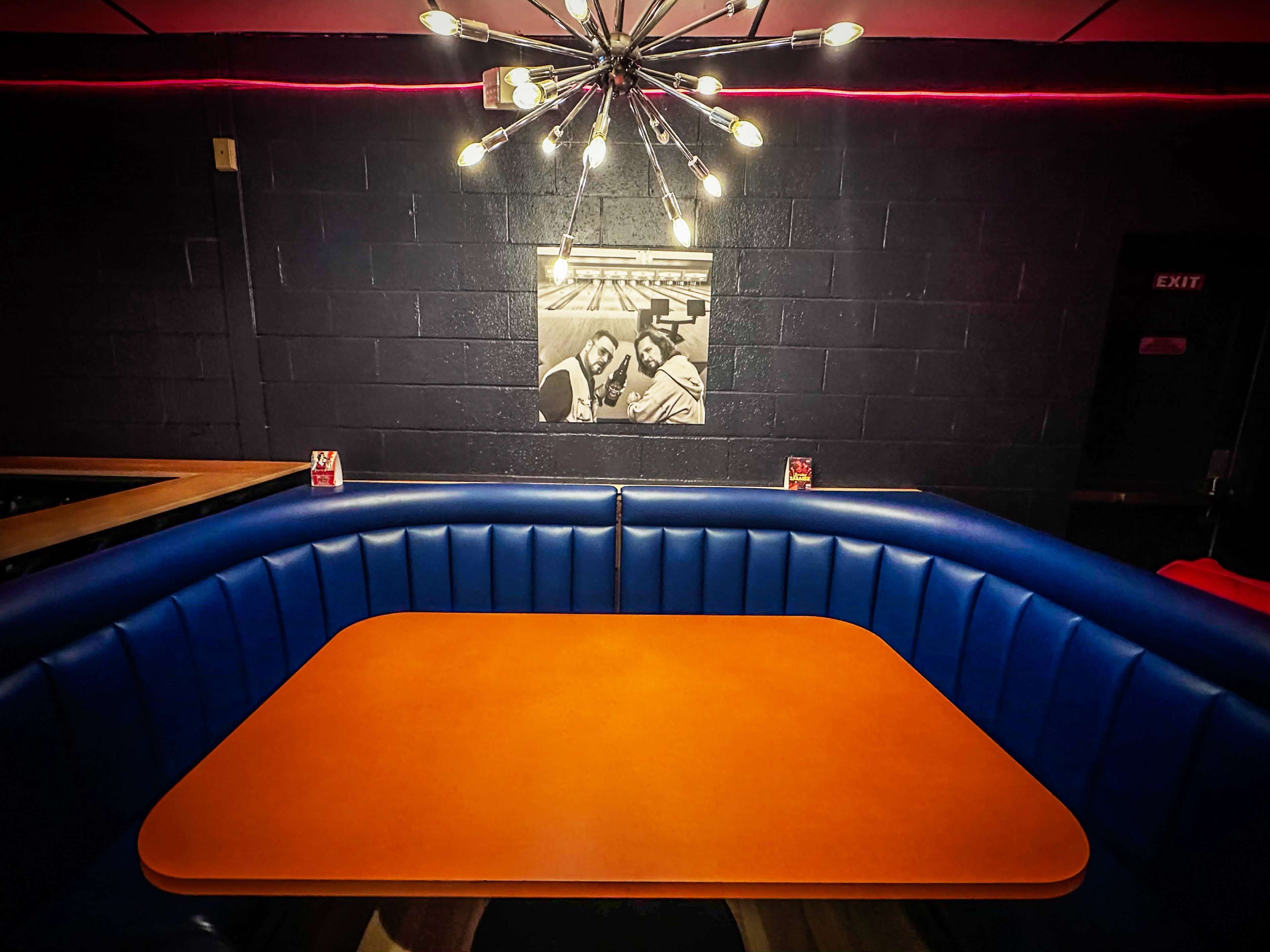 A blue leather booth with an orange table is illuminated by a starburst chandelier, with a black-and-white photograph on the wall behind.