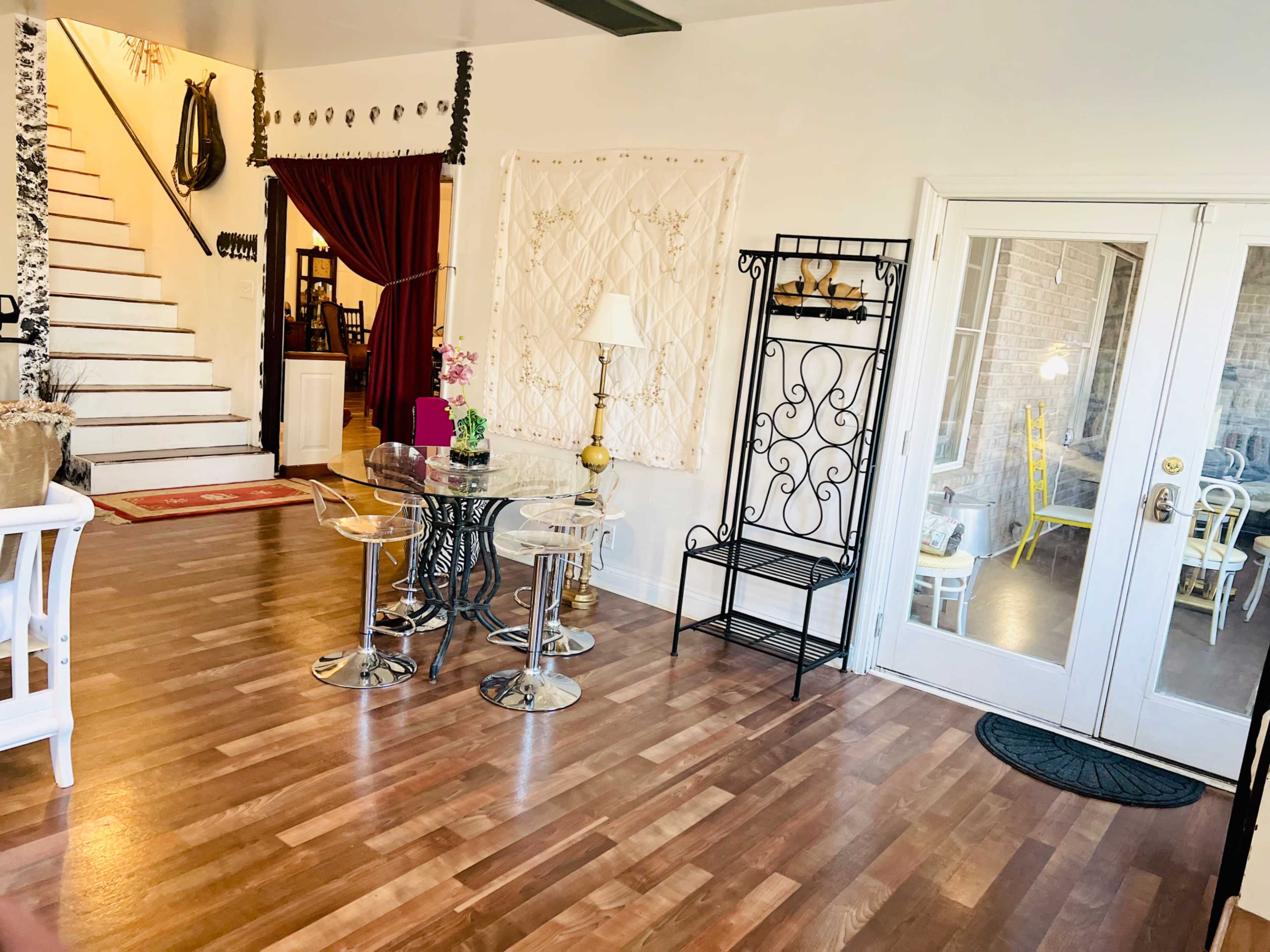 A bright indoor space featuring a dining area with a glass table and chairs, a staircase leading to a second level, and an entrance to a sunlit room through glass doors.