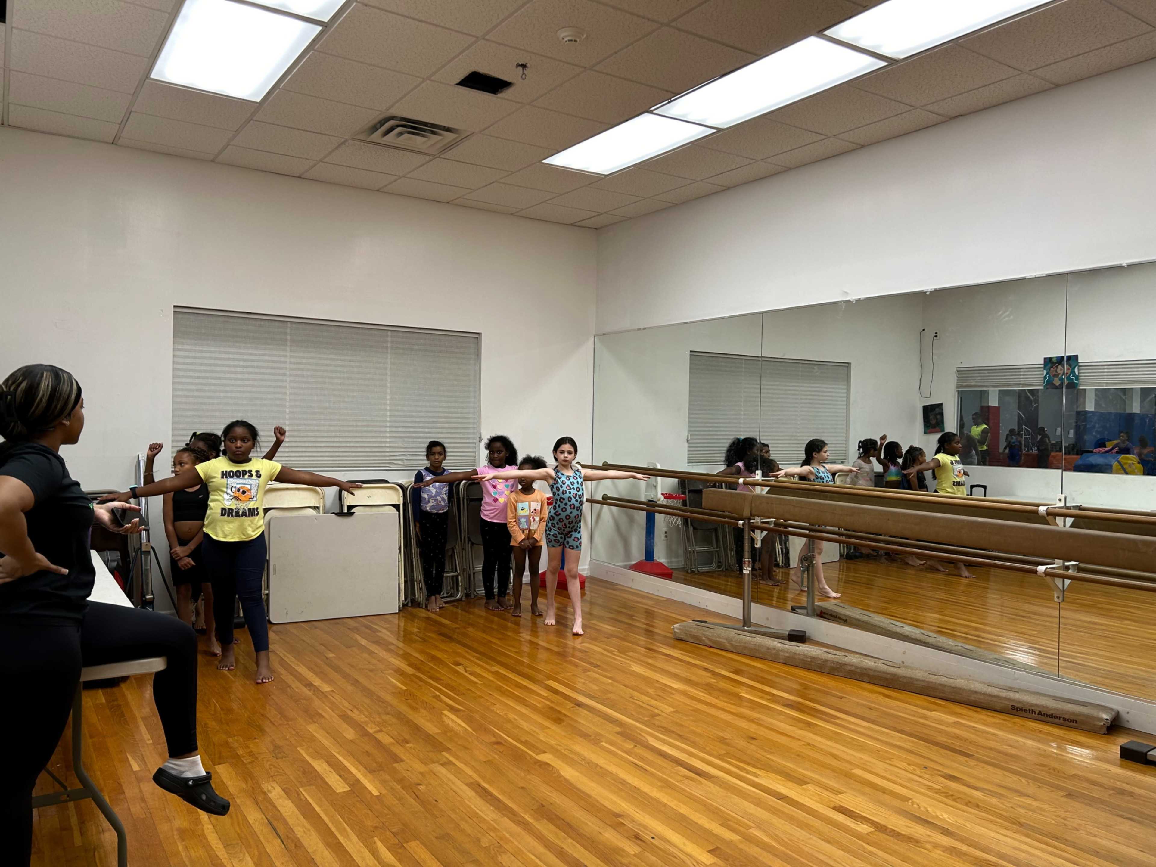 Several children stand at a ballet barre in a dance studio with mirrors on the wall, while an instructor looks on.