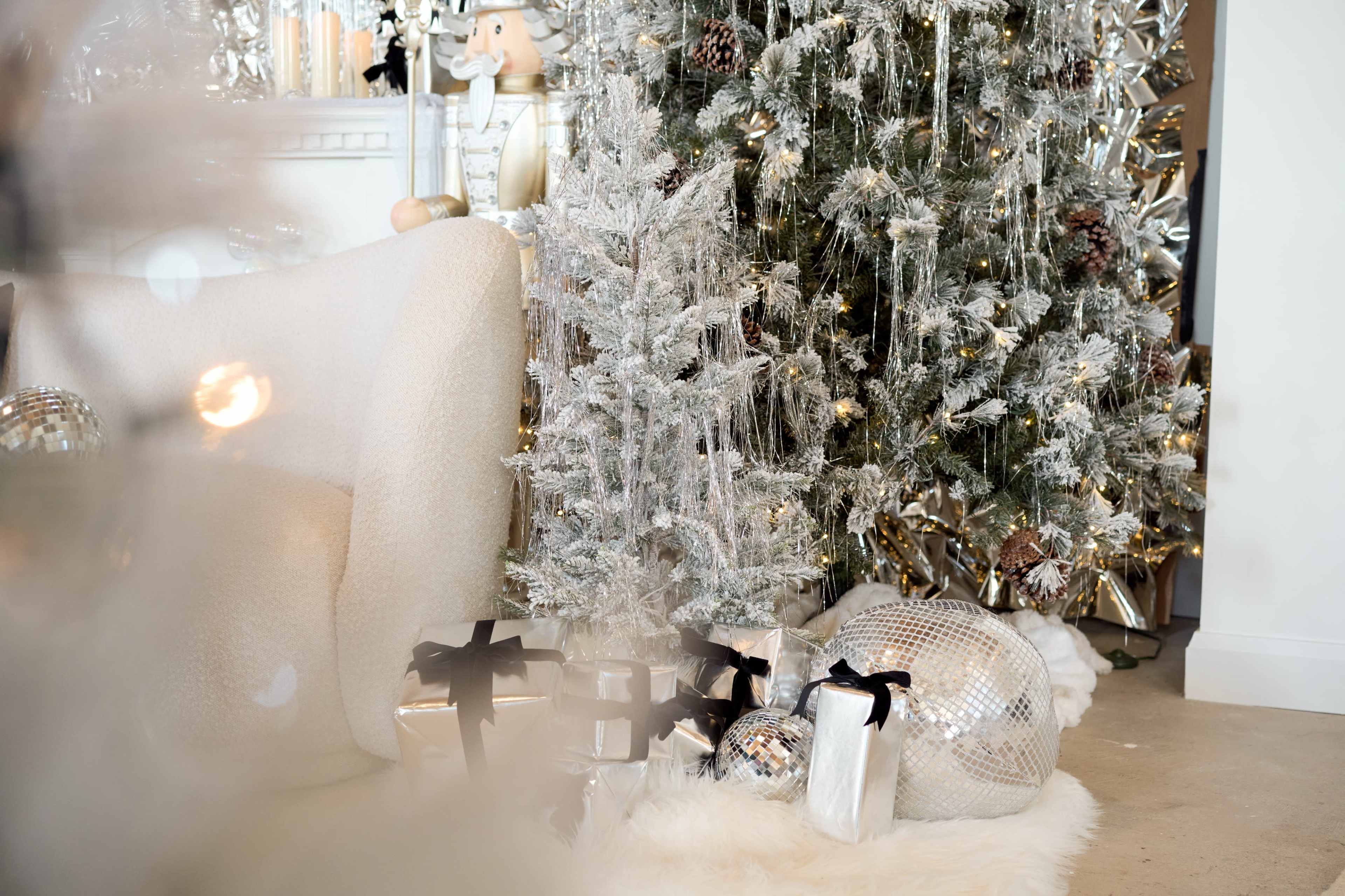 A silver Christmas tree adorned with decorations stands beside wrapped gifts on a plush white rug in a cozy living space.