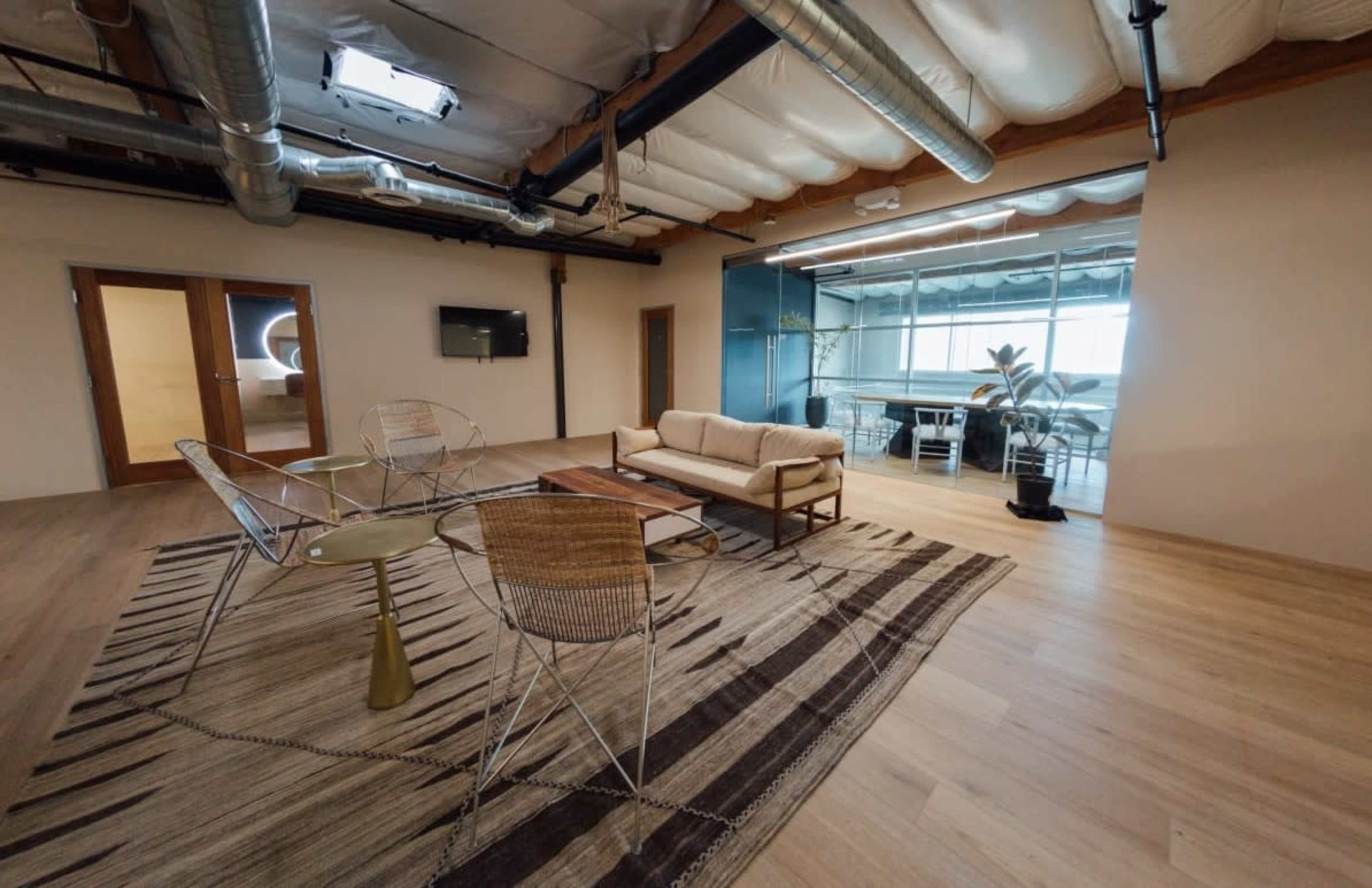 The image shows a modern office space with a seating area consisting of a sofa, a coffee table, and chairs on a patterned rug, along with large windows and potted plants in the background.